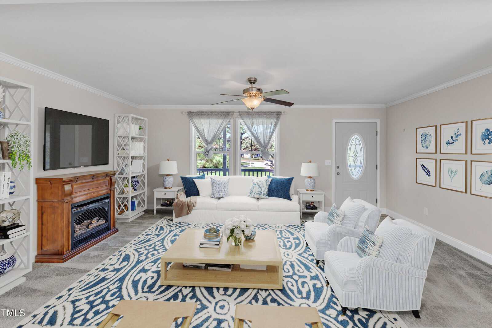 A brightly lit living room with a large white sofa, two armchairs, a wooden coffee table, and a plush blue-and-white patterned rug. A fireplace and TV stand are on the left, and framed artwork decorates the beige walls. Large windows have sheer curtains.
