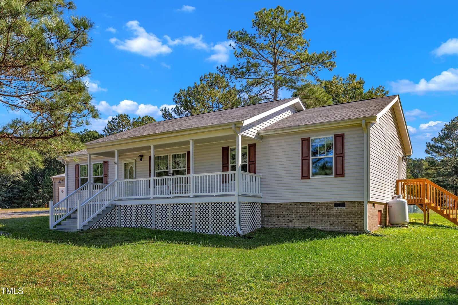 Nestled at 87 Stonebridge Lane, this charming single-story house in Henderson features a white exterior and a welcoming covered front porch. A wooden staircase leads up to the porch, flanked by large windows, lush green grass, and trees under a picturesque blue sky with clouds.