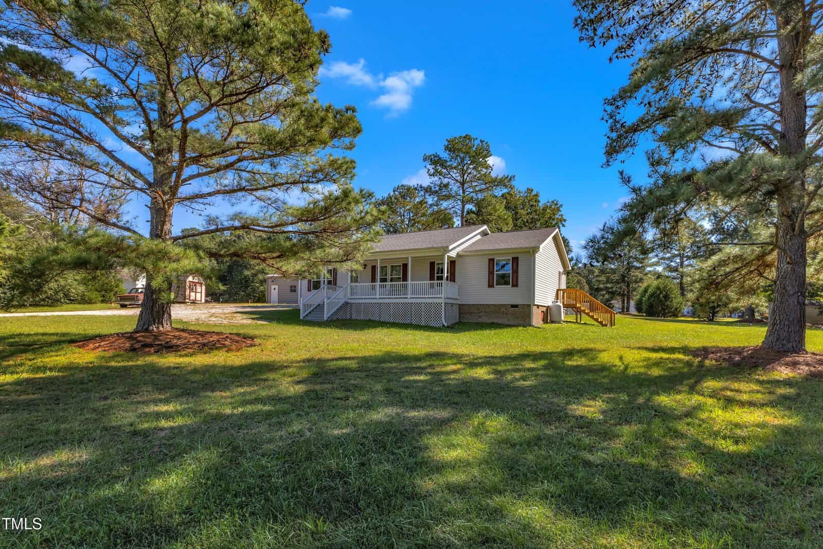 A white house with a spacious front porch is surrounded by green grass and tall trees. The sky is clear and blue, creating a peaceful, sunny atmosphere. The house has a sloped roof and is set back from a driveway.
