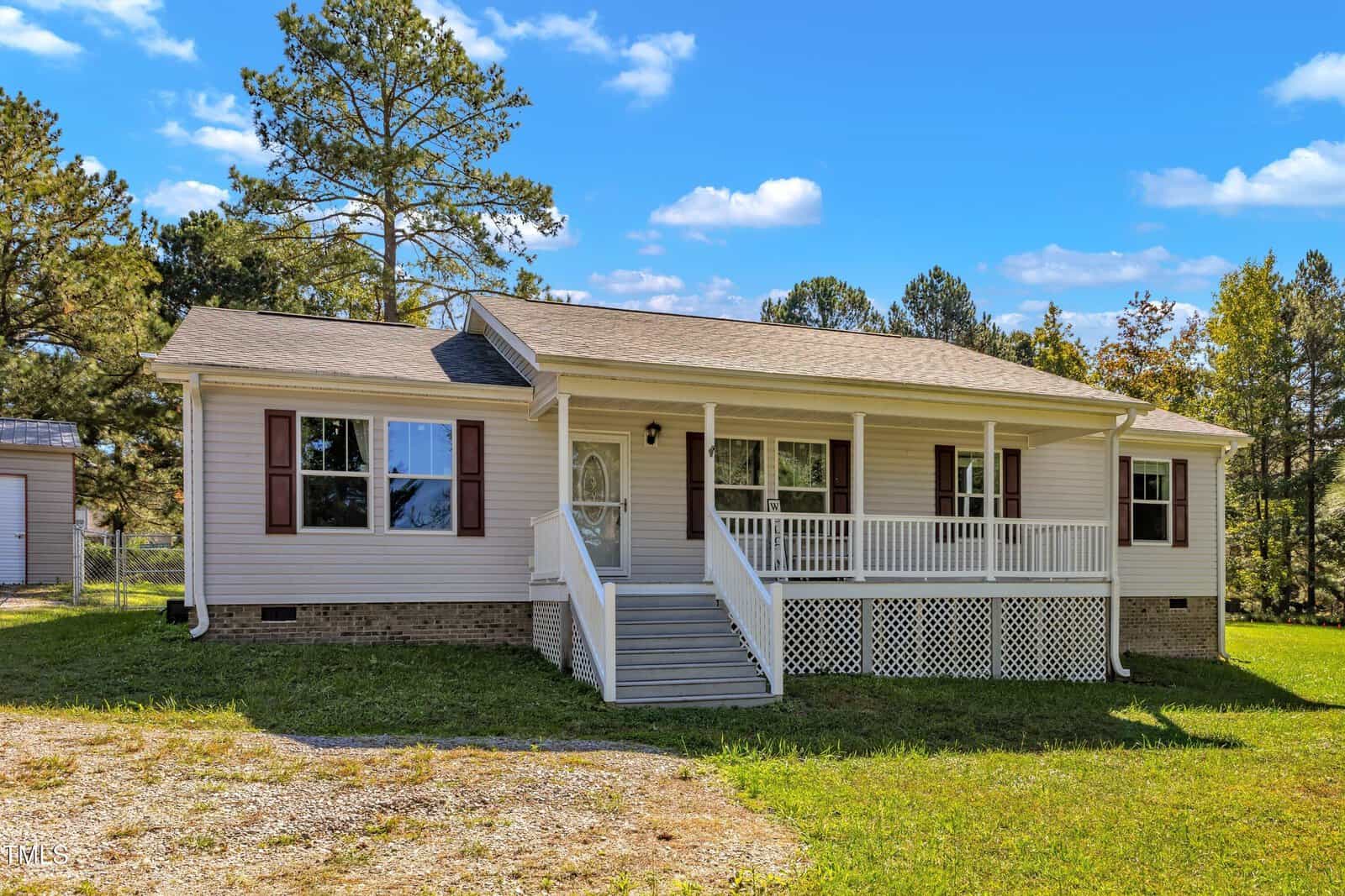 A single-story house with a light beige exterior, white trim, and a small front porch with railings. The yard is grassy, and trees are in the background. The sky is blue with clouds.