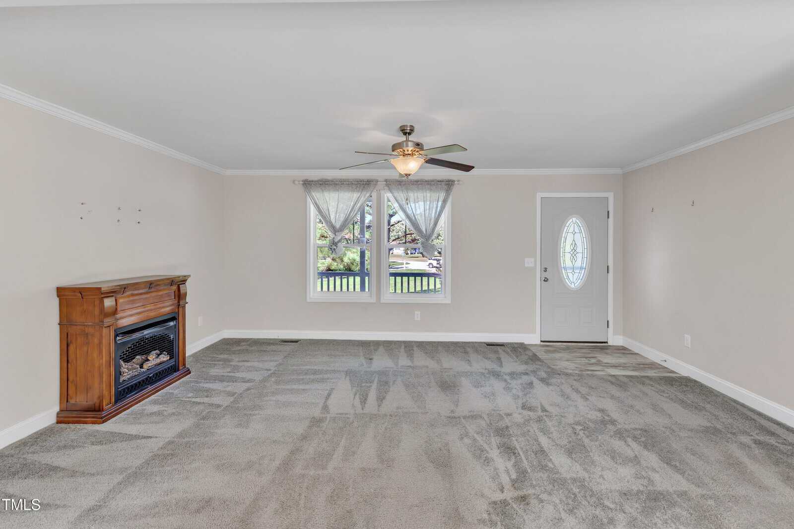 A spacious living room with light-colored walls and carpeted floor. It features a wooden fireplace on the left, a ceiling fan, a window with lace curtains, and a glass-paneled front door on the right. The room is bright and airy.