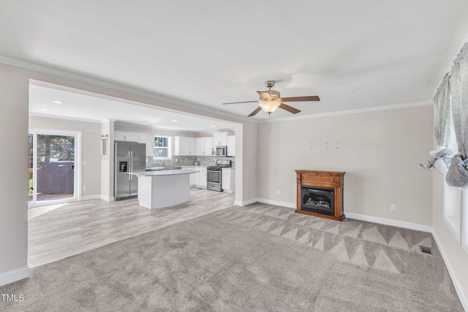A spacious living room with beige carpet and a ceiling fan. The room features a decorative wooden electric fireplace. An open layout leads to a modern kitchen with a large island, stainless steel appliances, and white cabinetry. Natural light fills the space.
