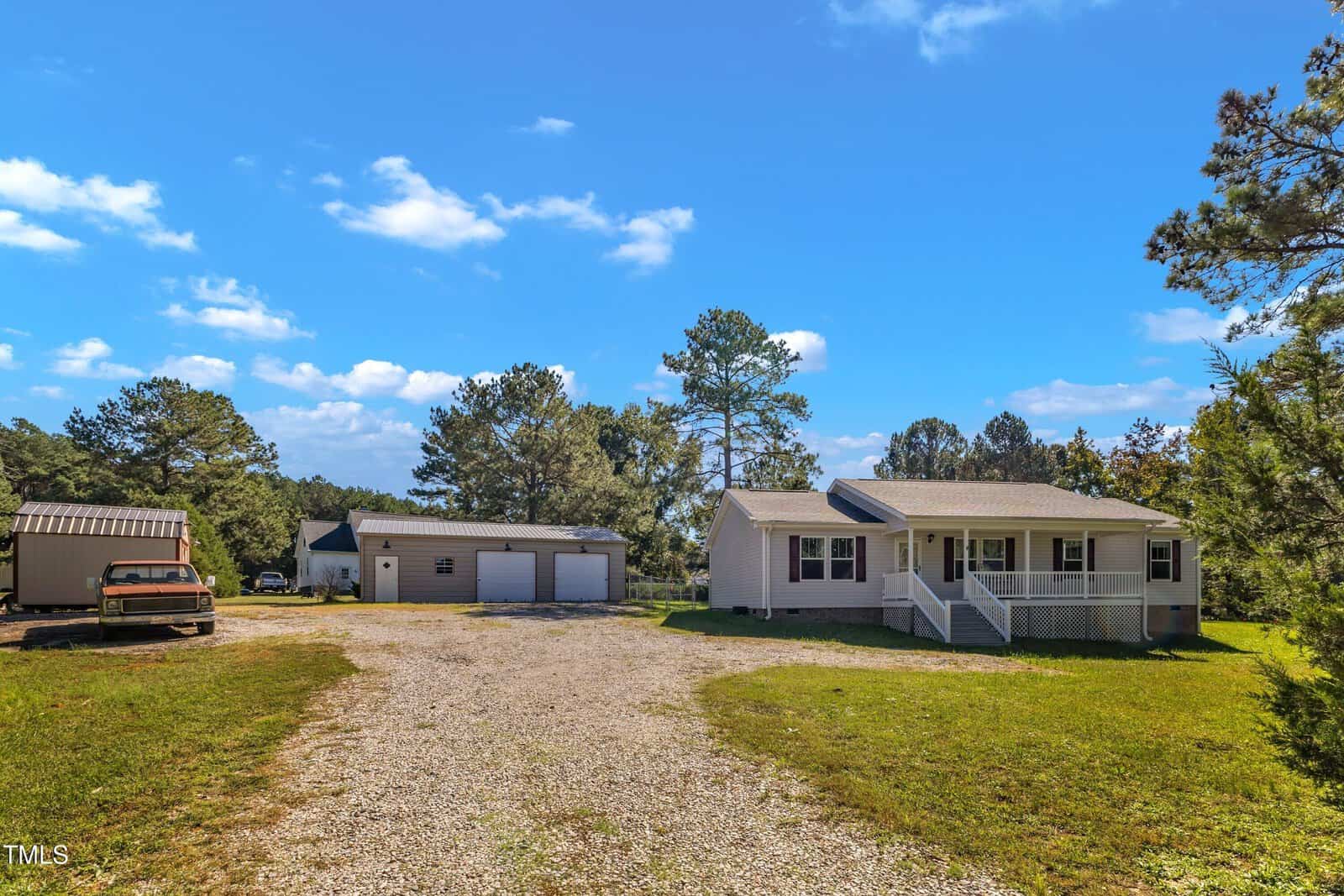 A white, single-story house with a front porch is situated on a grassy lot. There is a gravel driveway leading to a detached garage and an old truck on the left. Surrounding the property are trees under a bright blue sky with a few clouds.