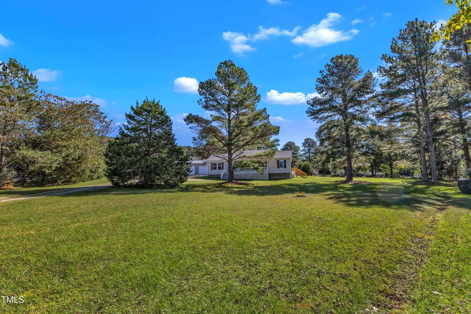 A single-story house with a light exterior is surrounded by green grass and trees under a blue sky. The landscape includes a spacious lawn and several tall pine trees.
