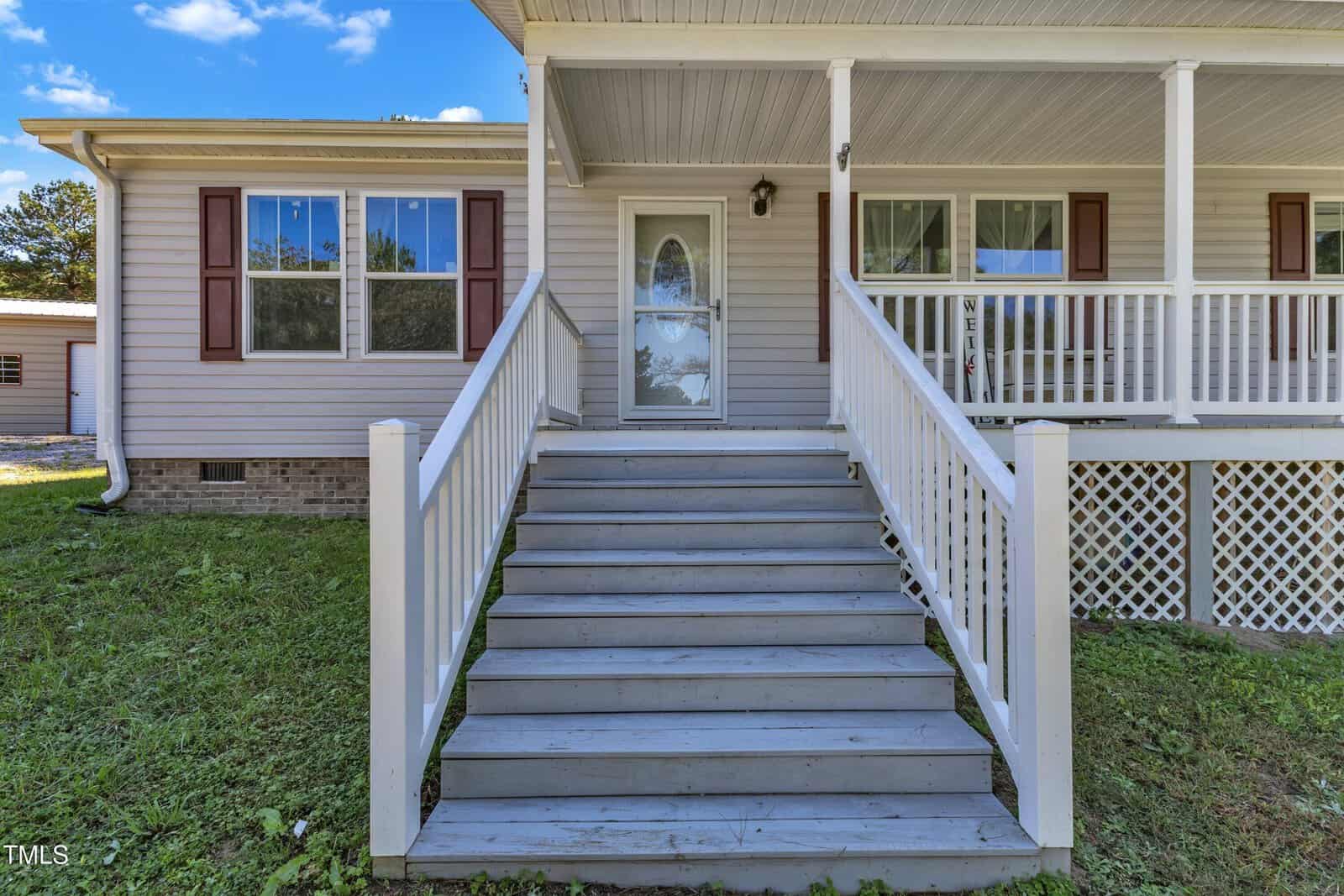 A light gray house with a wooden porch and white railings is shown. The front features a staircase leading to a doorway with a glass door. The porch is covered by a roof, and the yard is grassy.