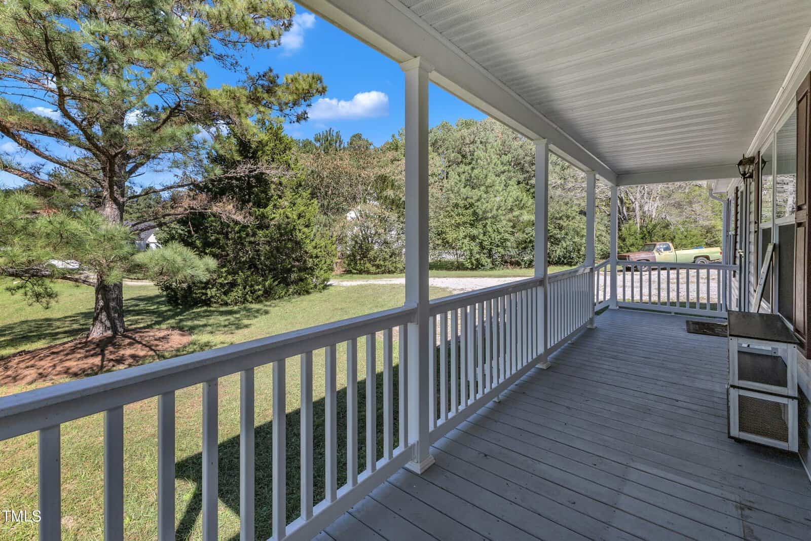 A spacious, covered wooden porch with white railings overlooks a green lawn and trees under a clear blue sky. A small bench and a doormat are near the entrance. The setting is peaceful and serene.