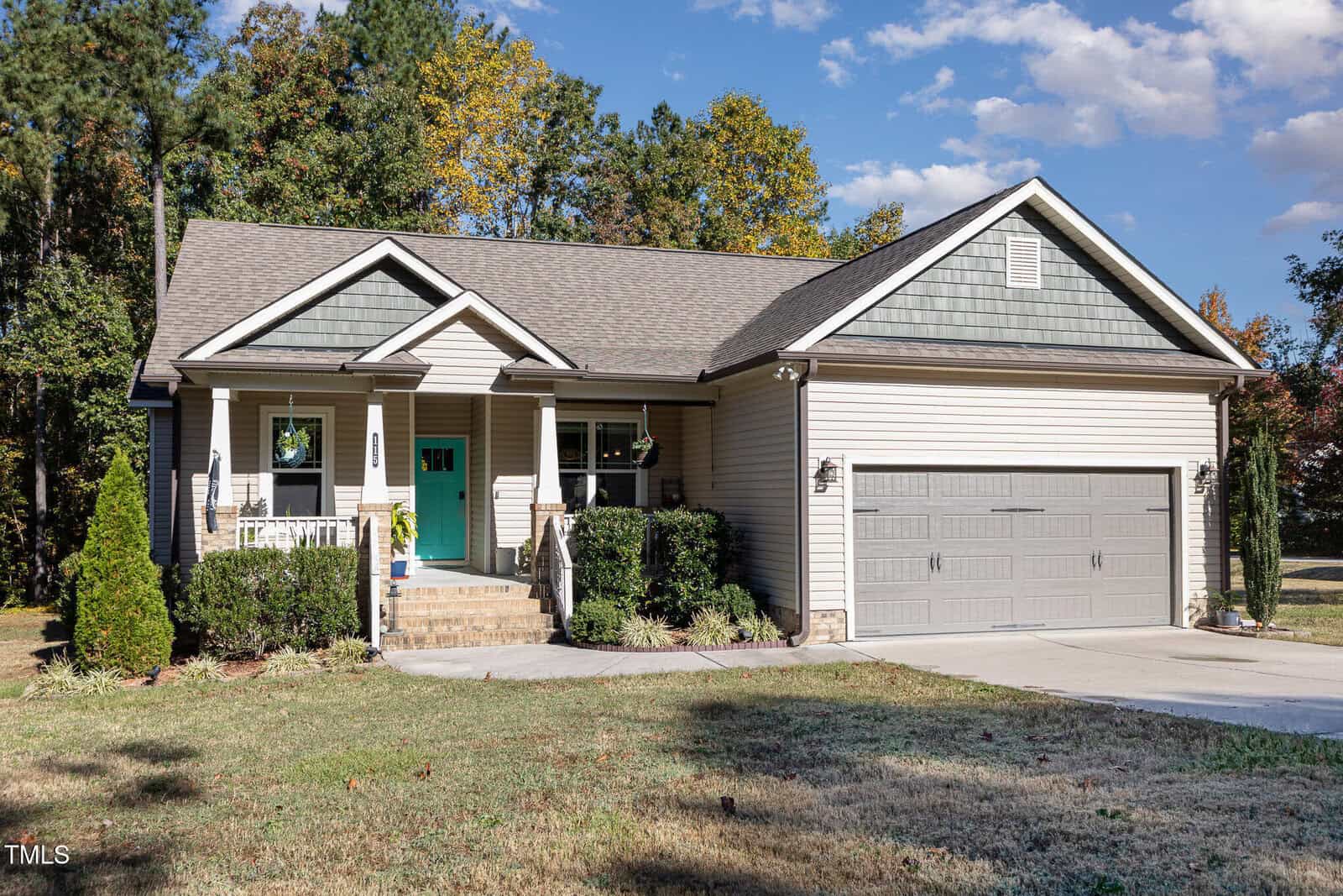 A single-story suburban house with gray siding, a green front door, a small covered porch, and a two-car garage. The front yard is grassy with bushes and trees in the background under a partly cloudy sky.
