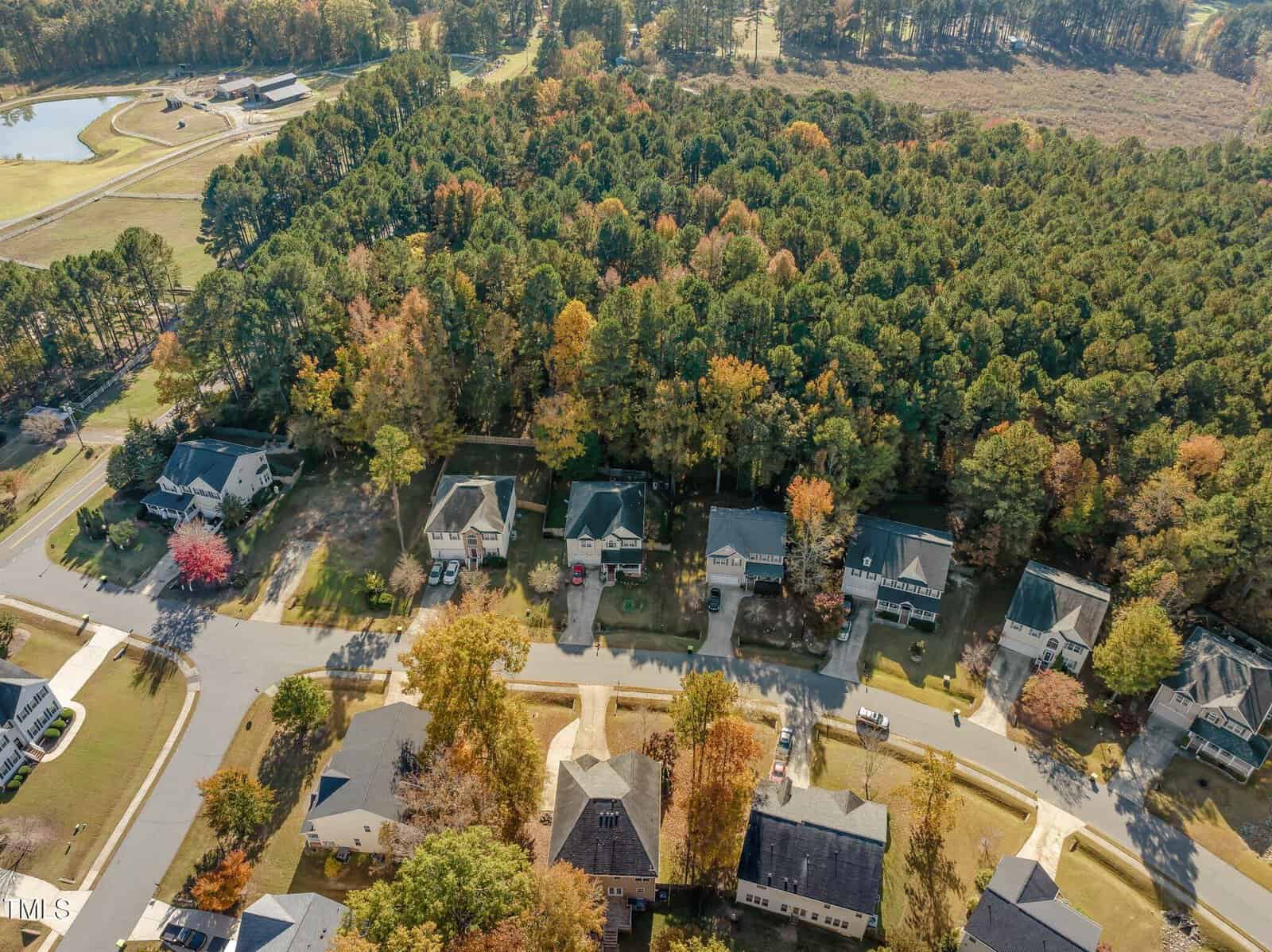 Aerial view of a suburban neighborhood with houses lined up along winding streets. There is a dense forested area in the background, and some trees show fall colors. A small pond is visible in the top left corner.