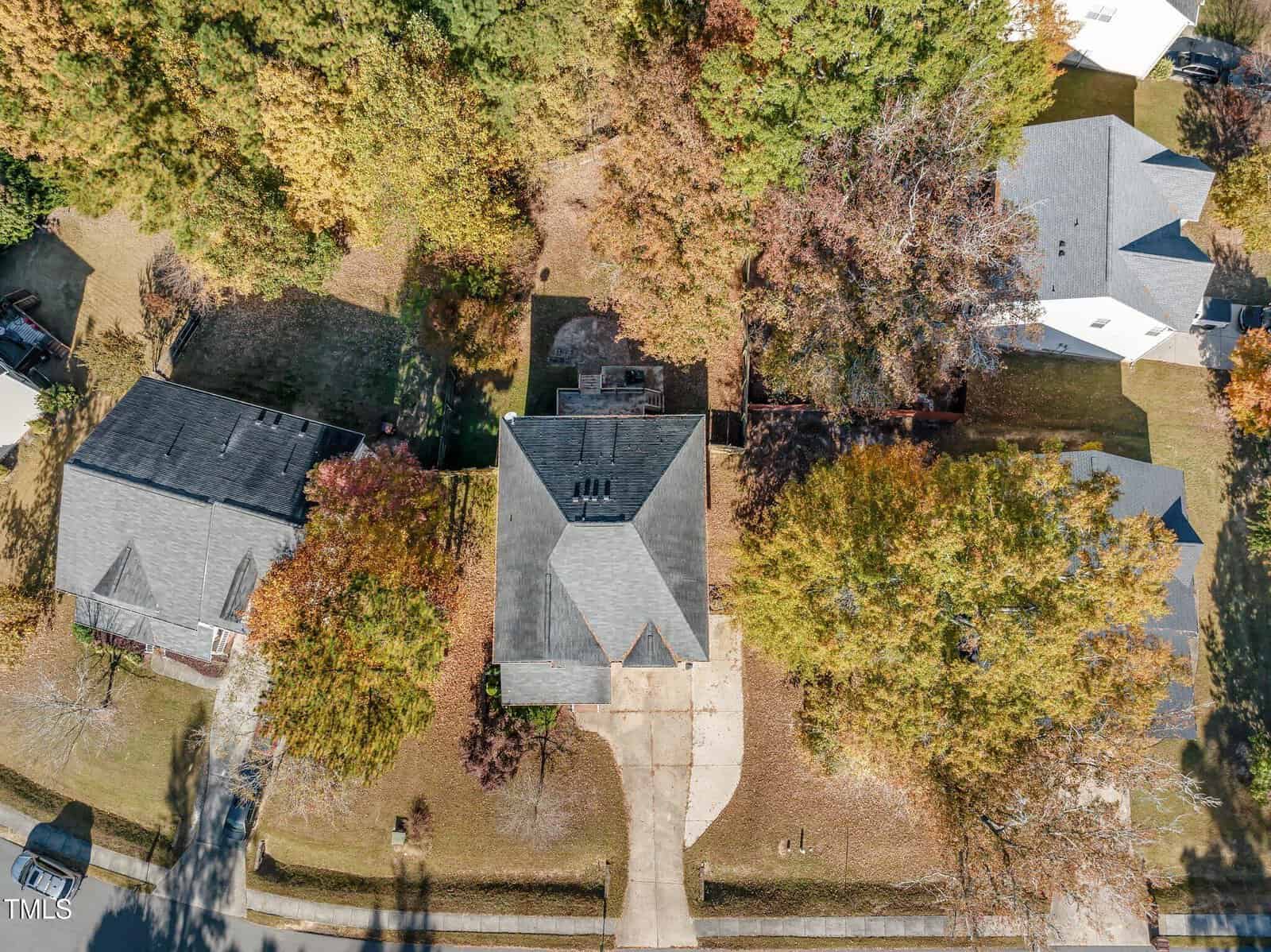 Aerial view of a suburban neighborhood showing a single-story house with a gray roof, surrounded by trees. The house has a two-car driveway, a front yard, and is flanked by similar homes on either side. The trees display autumn foliage.