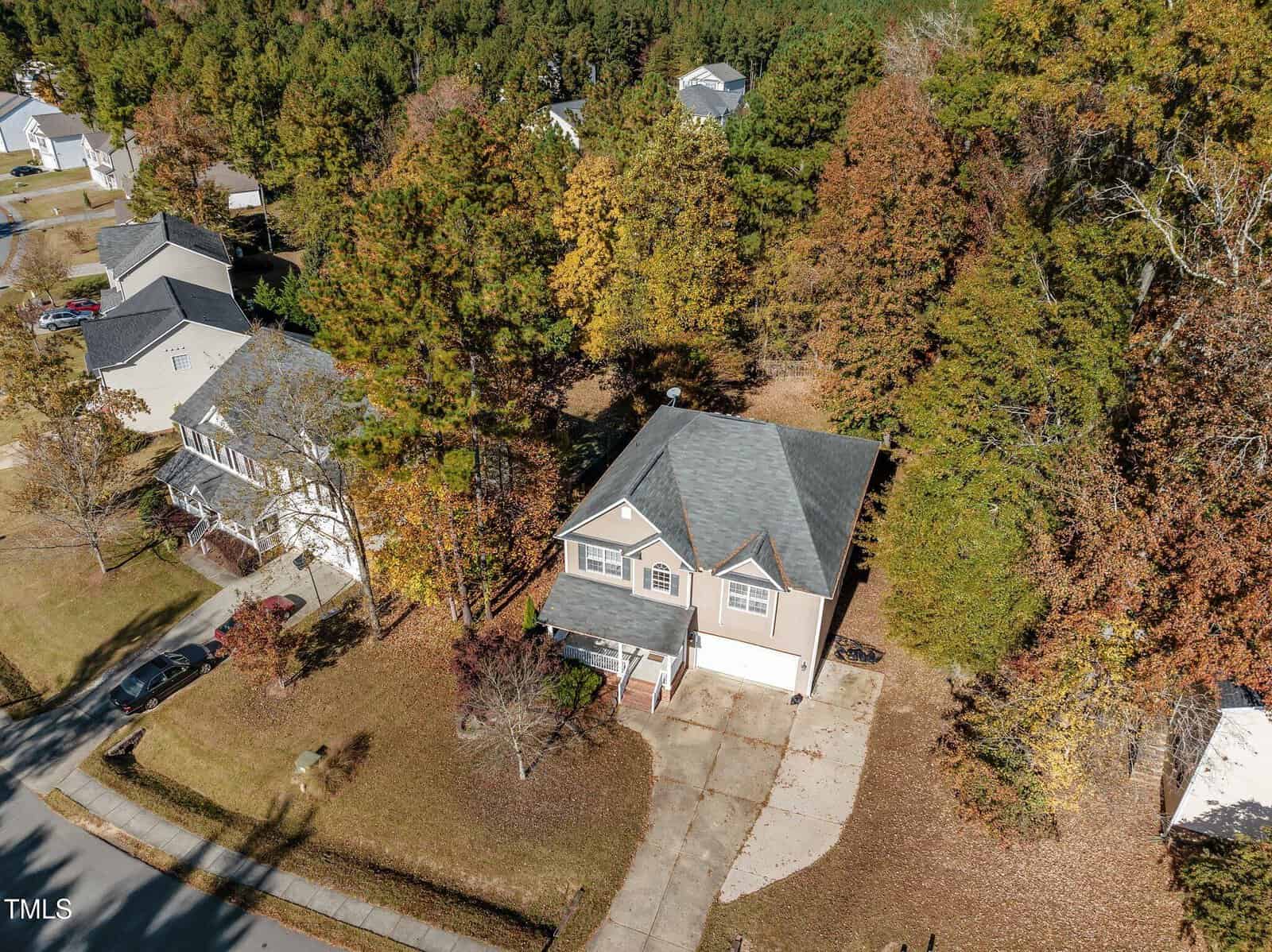 Aerial view of a suburban neighborhood with several houses surrounded by trees. The main focus is a two-story house with a gray roof and a driveway, set among autumn foliage. Nearby homes and tree-lined streets are visible.