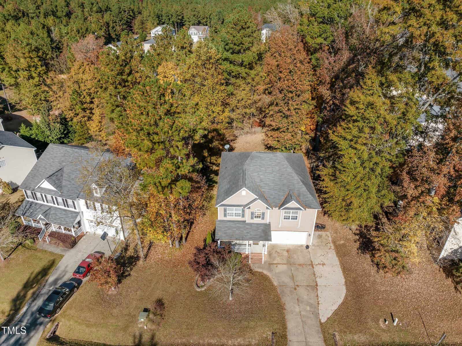 Aerial view of a suburban neighborhood with two houses surrounded by deciduous trees in autumn. The main house has a driveway extending to the street, and the area shows fall foliage with brown and orange leaves.