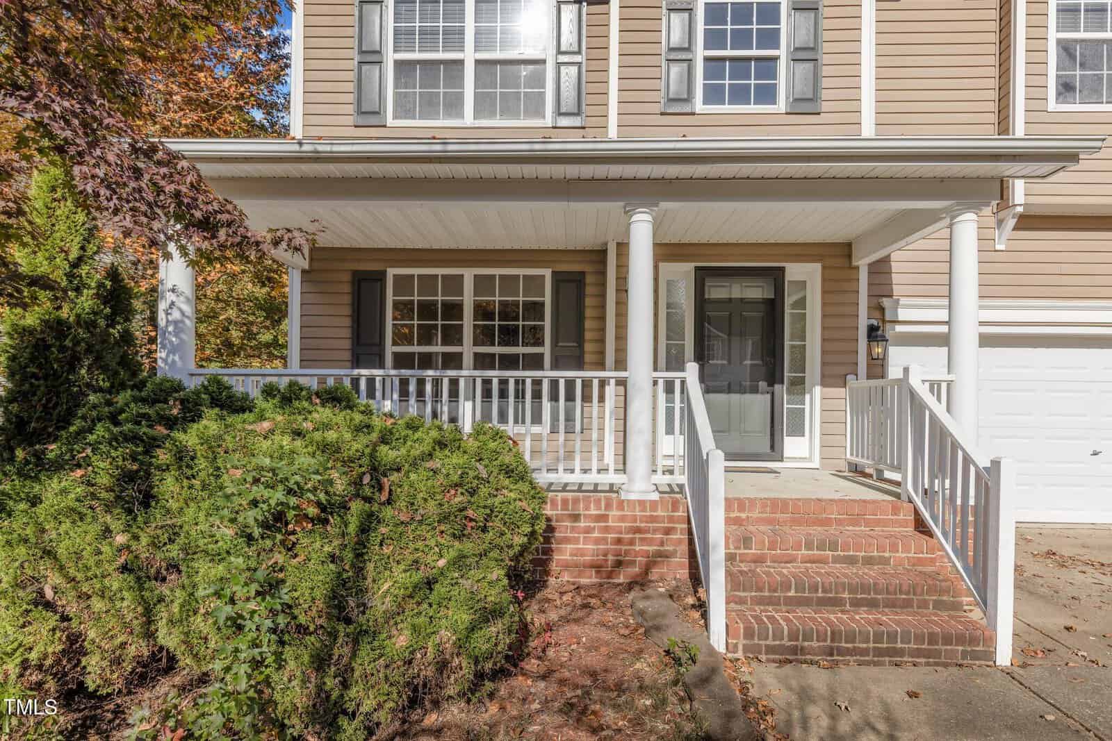 A two-story beige house with white trim features a covered front porch supported by white columns. It has a glass front door and several windows. The porch is accessible by brick steps, and there is a garage door on the right. Green shrubs line the front.