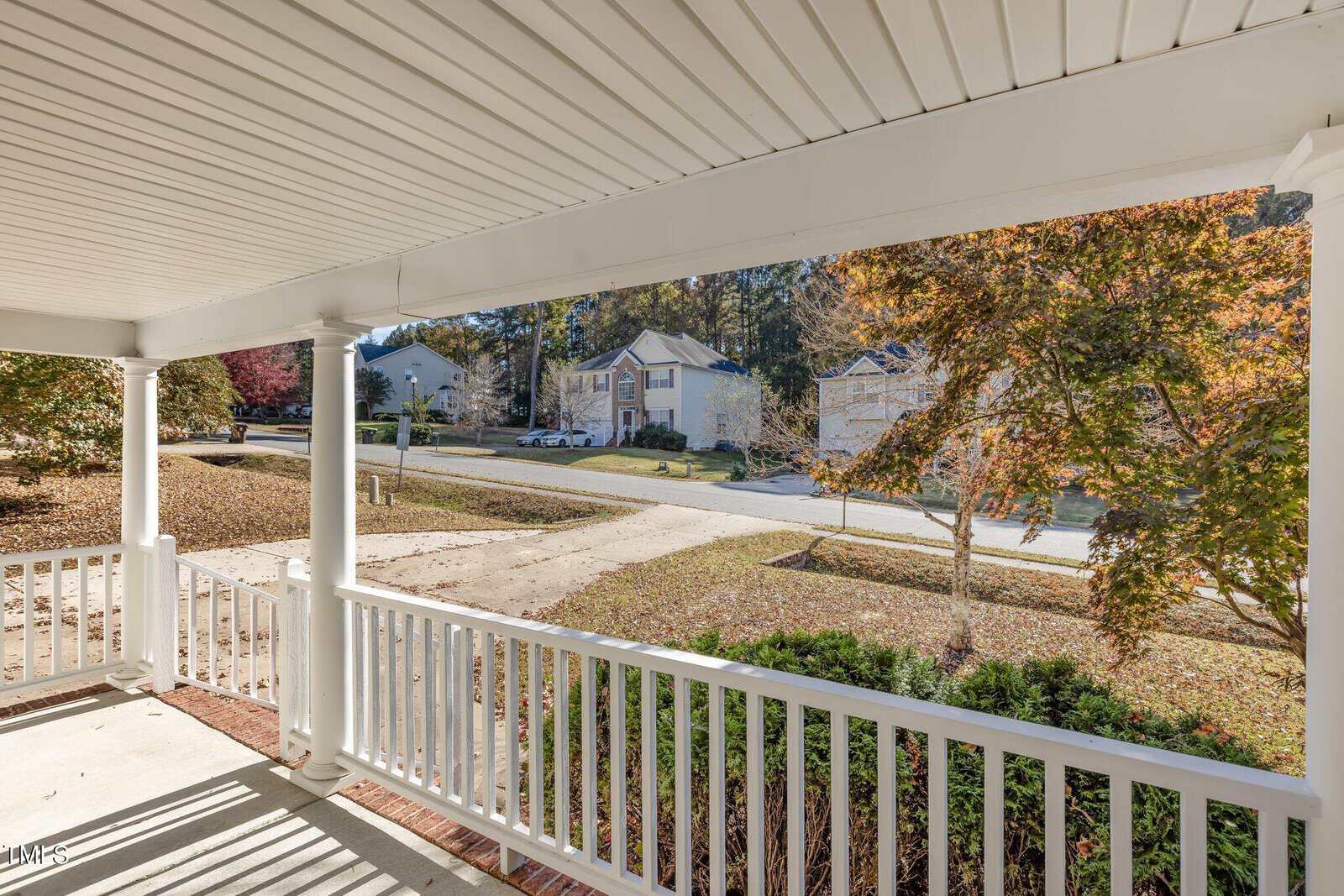 View from a covered porch with white railings and columns, overlooking a suburban street lined with trees and houses. The ground is covered with fallen leaves, and the sky is clear.