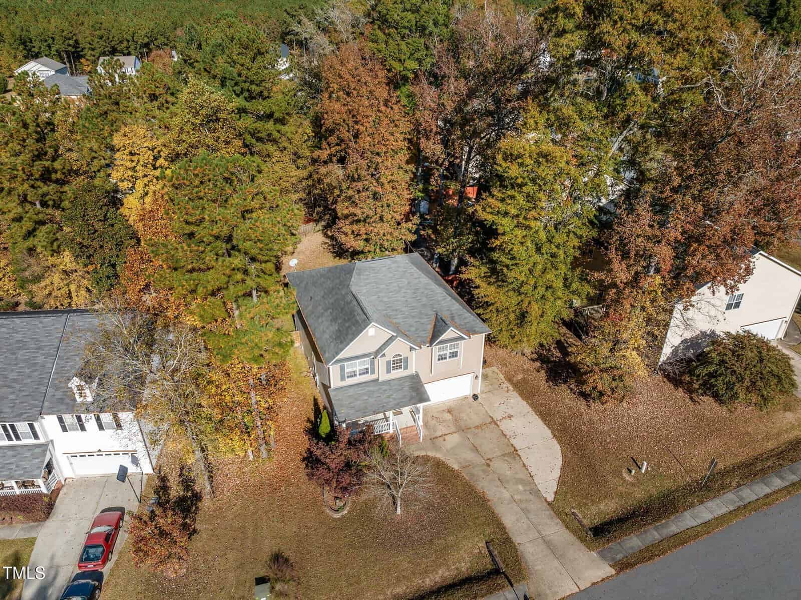 Aerial view of a suburban house with a gray roof and beige exterior, surrounded by tall trees with autumn foliage. The property includes a driveway and is located in a neighborhood with similar homes.