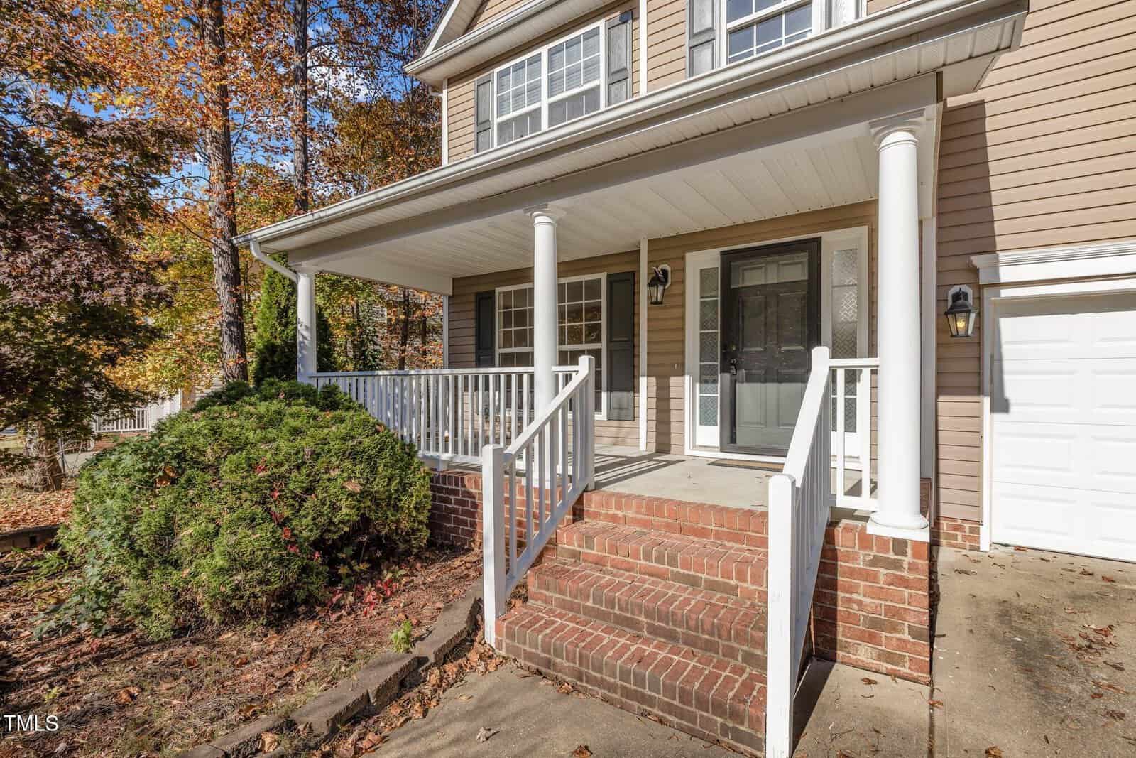 A house with a beige exterior features a covered front porch supported by white columns. Brick steps lead up to the porch, which is bordered by a white railing. The background includes trees with autumn foliage.