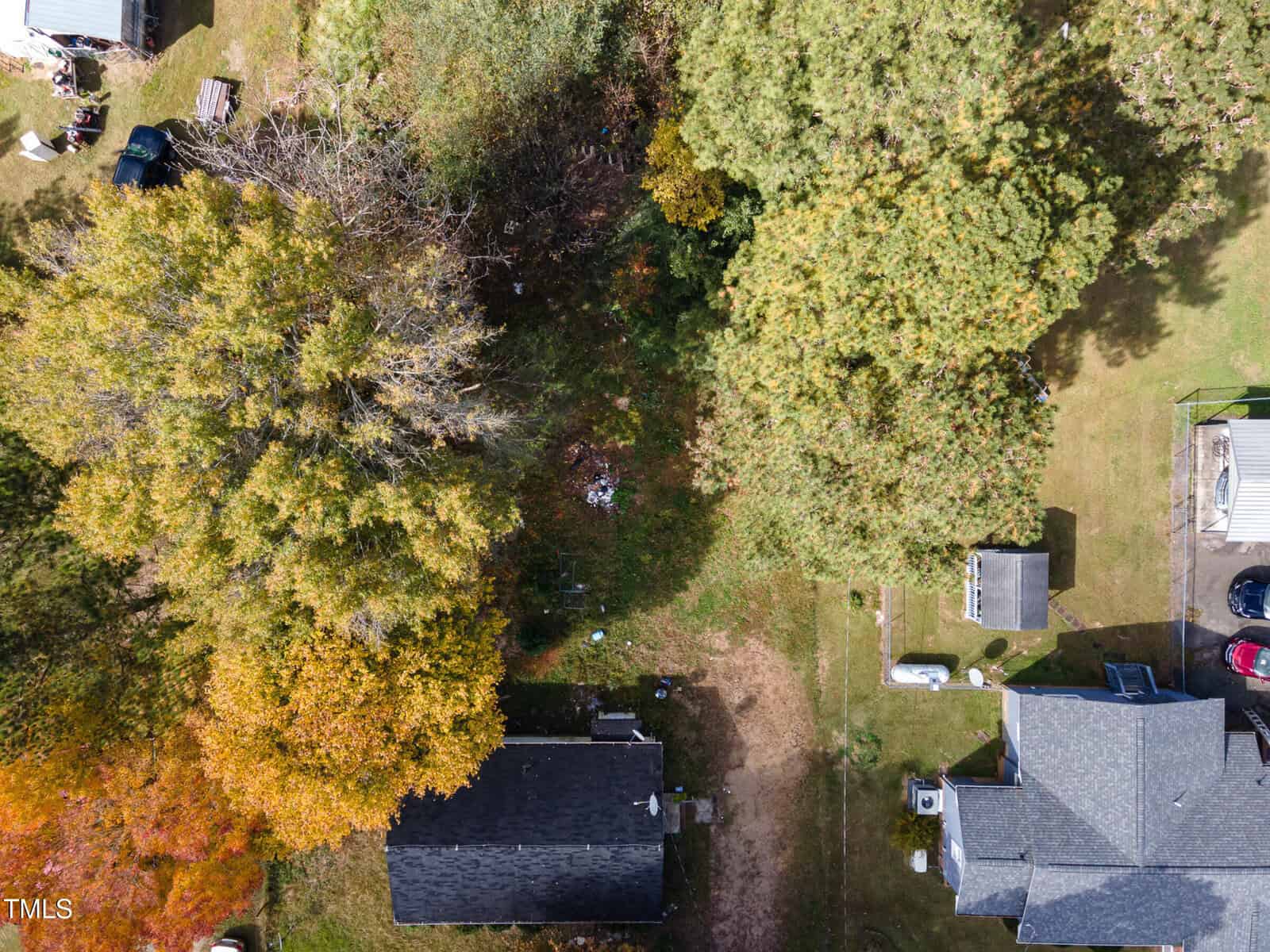 Aerial view of a residential area on 2065 Enon Road in Oxford, showcasing two houses enveloped by trees. The trees display autumn hues of green, yellow, and orange. Nearby, vehicles rest on a driveway amidst patches of grass and dirt.