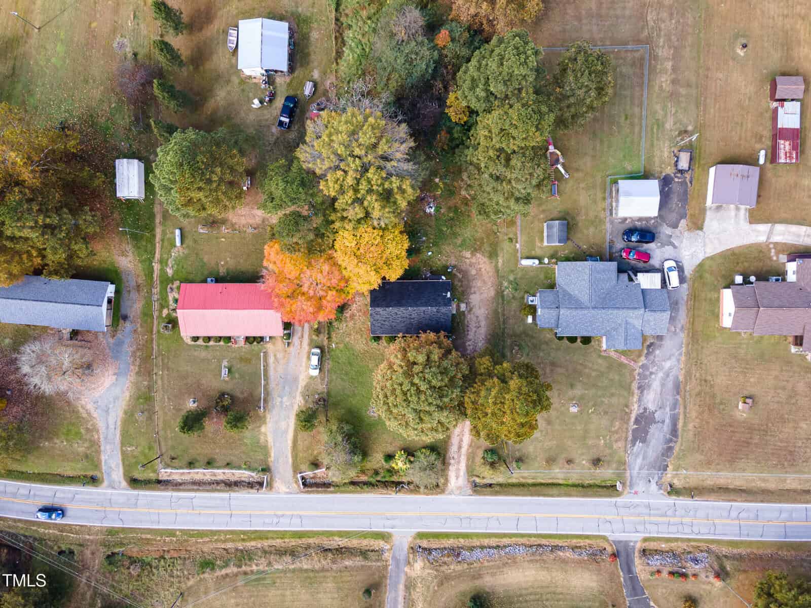 Aerial view of a rural neighborhood with five houses surrounded by trees. A road runs horizontally at the bottom of the image. One house is marked by vibrant autumn foliage. Cars are parked in driveways, and a small playground is visible.