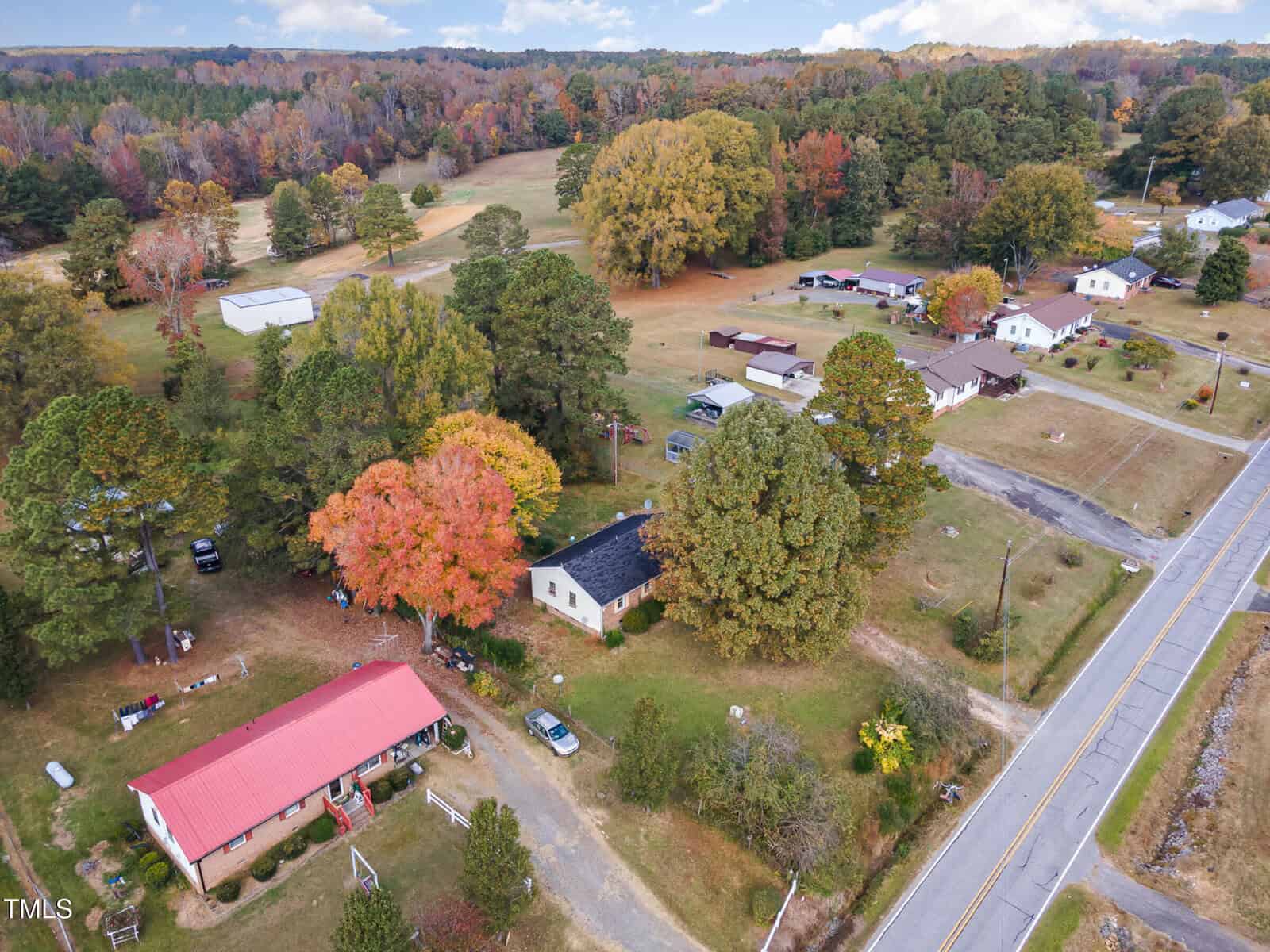 Aerial view of a rural landscape featuring scattered houses, trees with autumn foliage, open fields, and a road cutting through the area. The trees display vibrant shades of red, orange, and green, under a partly cloudy sky.