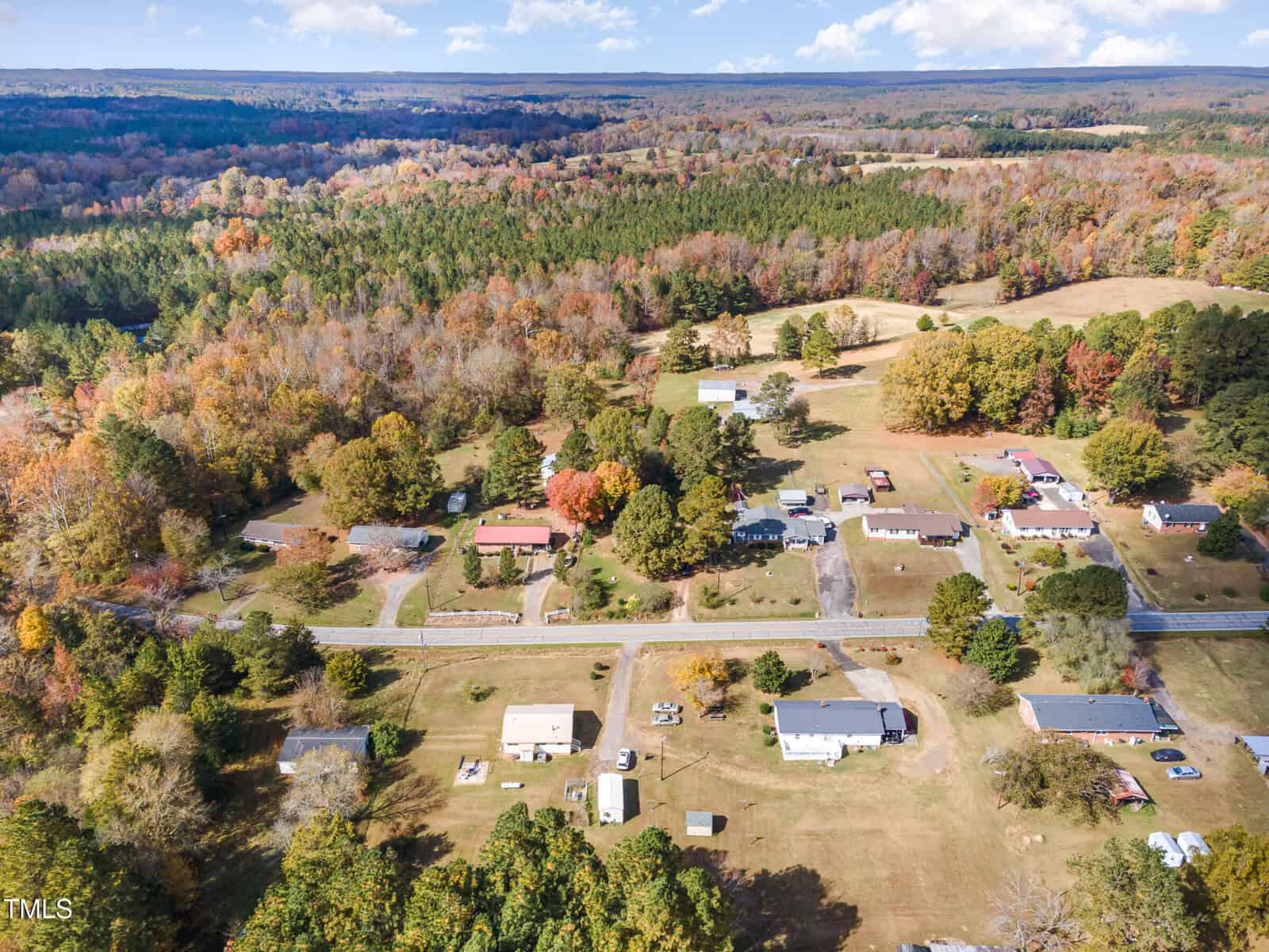 Aerial view of a rural landscape featuring a small residential area surrounded by fields and dense forests. The fall foliage creates a colorful tapestry of greens, oranges, and browns, with a clear sky overhead. A road runs through the residential area.