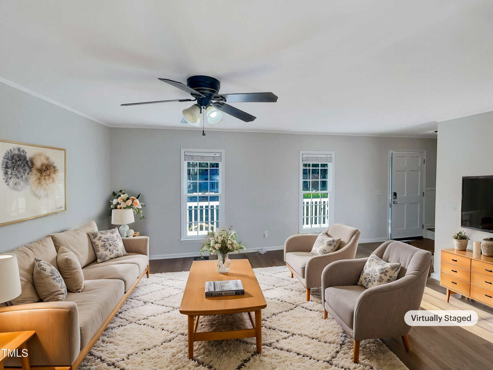 A virtually staged living room features a ceiling fan, two windows, and a door. The room includes a sofa, two armchairs, a coffee table, and a sideboard. Decor items such as a rug, plants, and wall art are present.