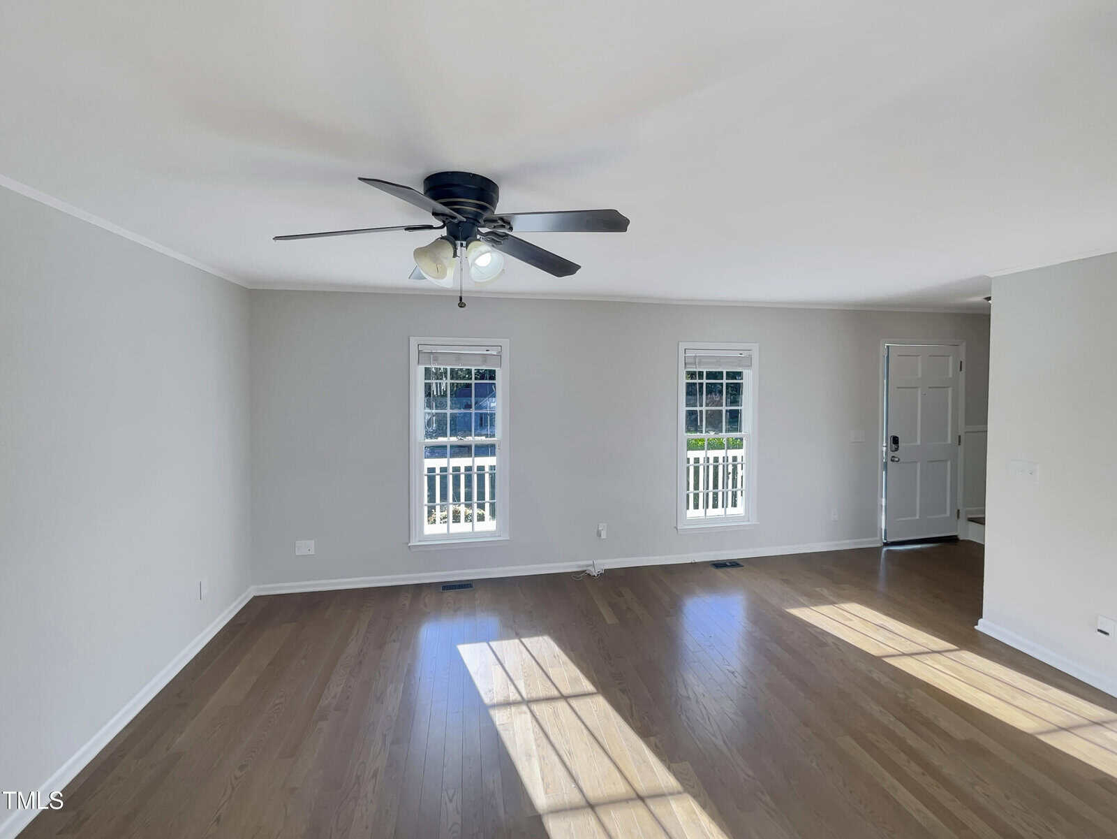 An empty room features polished hardwood floors and white walls. A ceiling fan with lights is centered above. Two windows and a door let in natural light, casting shadows on the floor.