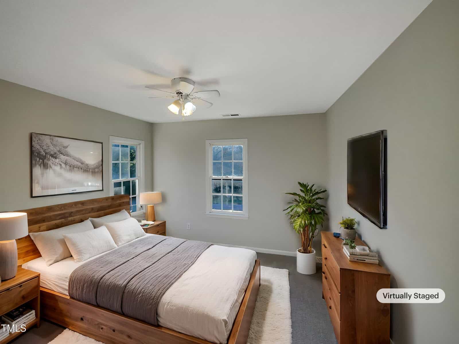 A bedroom with a wooden bed frame and gray bedding, two bedside tables with lamps, a flat-screen TV mounted on the wall, a dresser with a potted plant and decor, and a ceiling fan. The room is labeled "Virtually Staged.