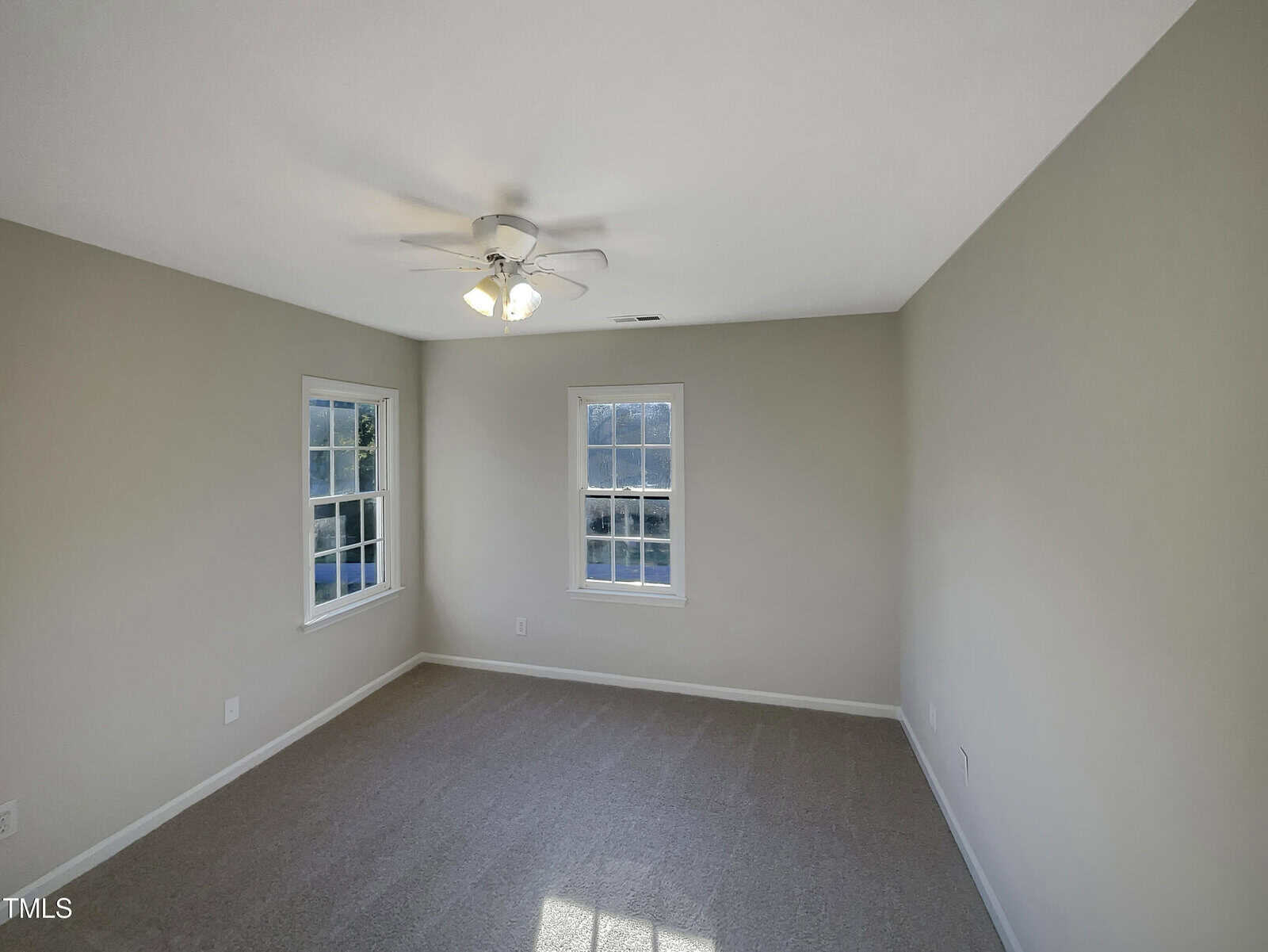 A vacant room with beige walls and a gray carpeted floor. There is a ceiling fan with lights, and three windows providing natural light.