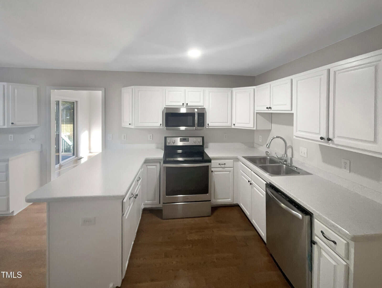 A modern kitchen with white cabinets and gray countertops. Features stainless steel appliances, including a refrigerator, stove, microwave, and dishwasher. Hardwood flooring is visible, and there is a windowed door leading outside.
