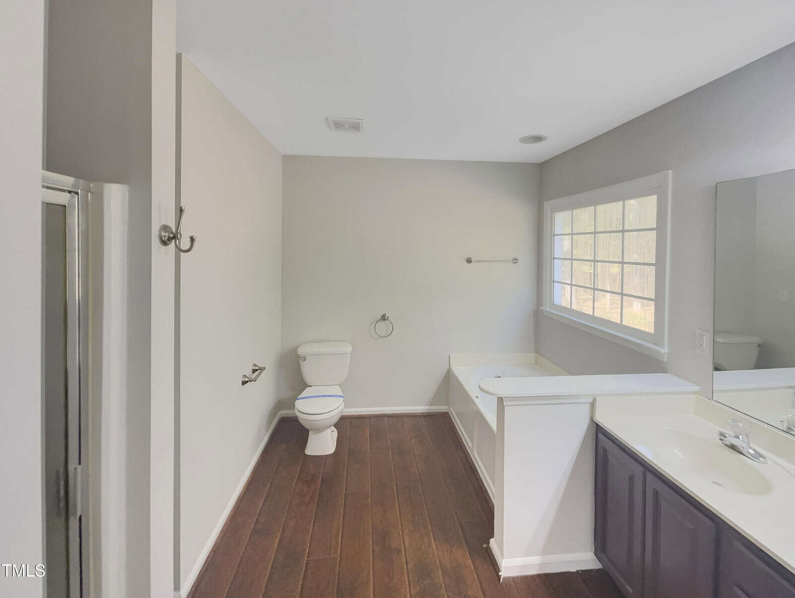 A bathroom with a toilet, a bathtub, and a glass-enclosed shower. There is a large window above the bathtub, a long countertop with a sink, and wooden flooring. The walls are painted in a light color.