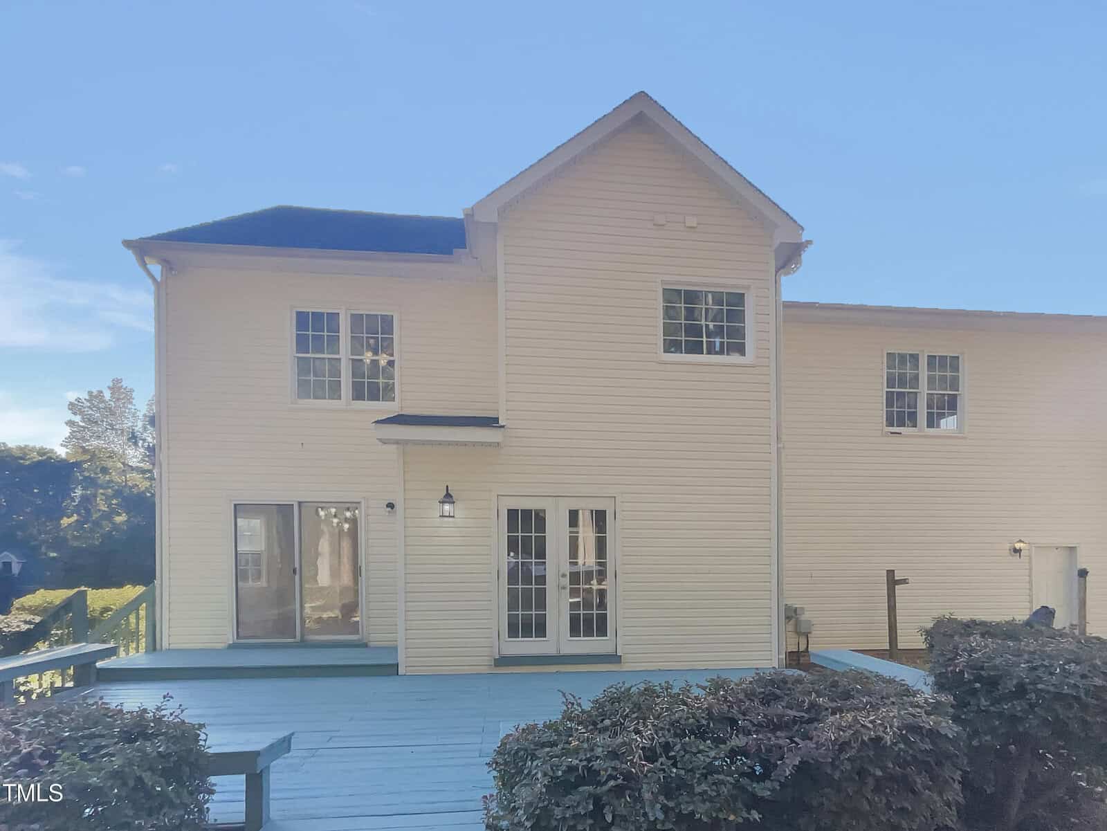 The image shows the back of a two-story house with light beige siding. There is a large blue painted wooden deck attached, surrounded by bushes. Multiple windows and a double glass door are visible on the house, and the sky is clear.