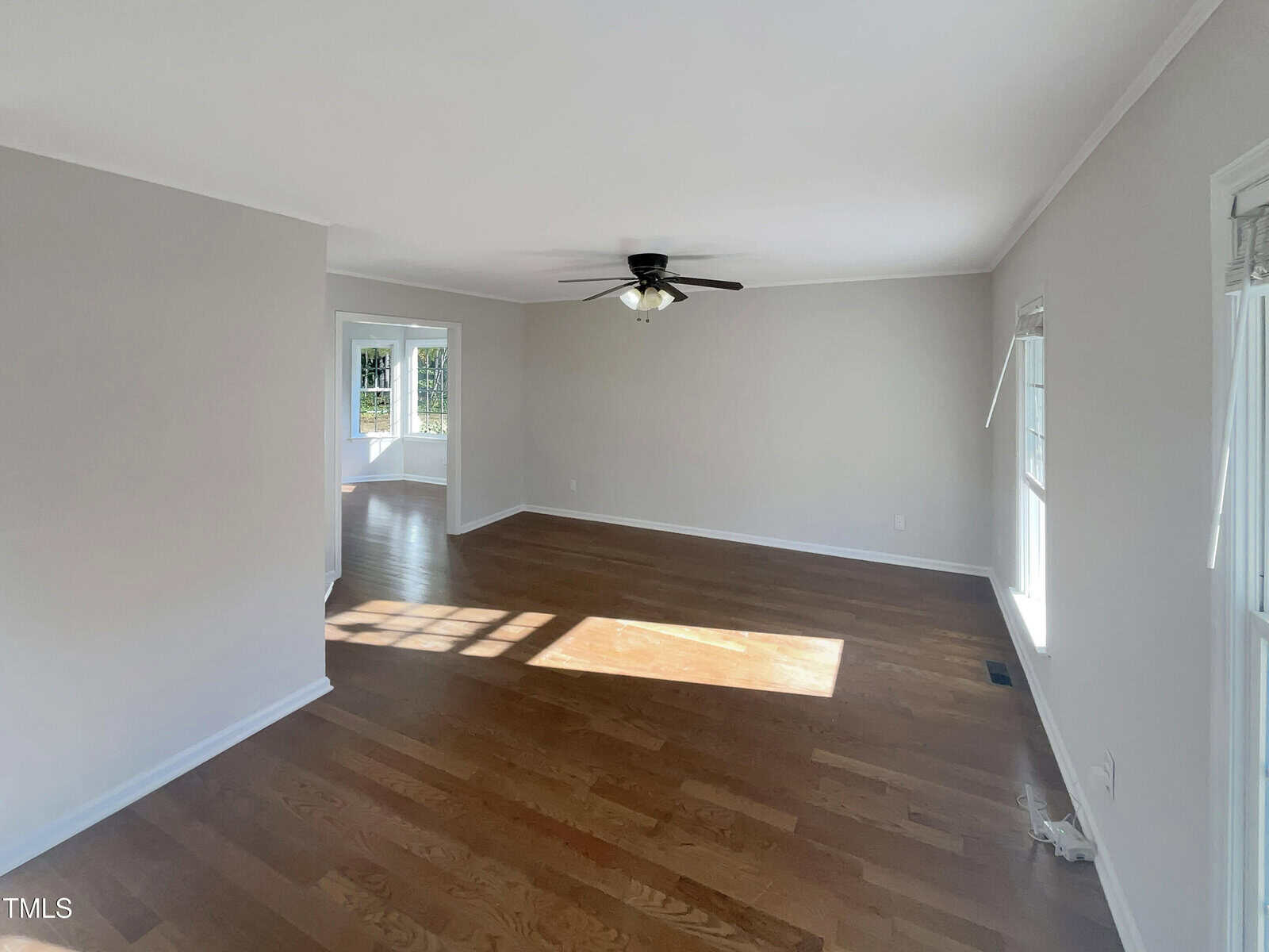 An empty living room with light brown hardwood flooring and beige walls. There is a ceiling fan with lights in the center. Sunlight streams through windows on the right, and an adjoining room is visible through an opening on the left.