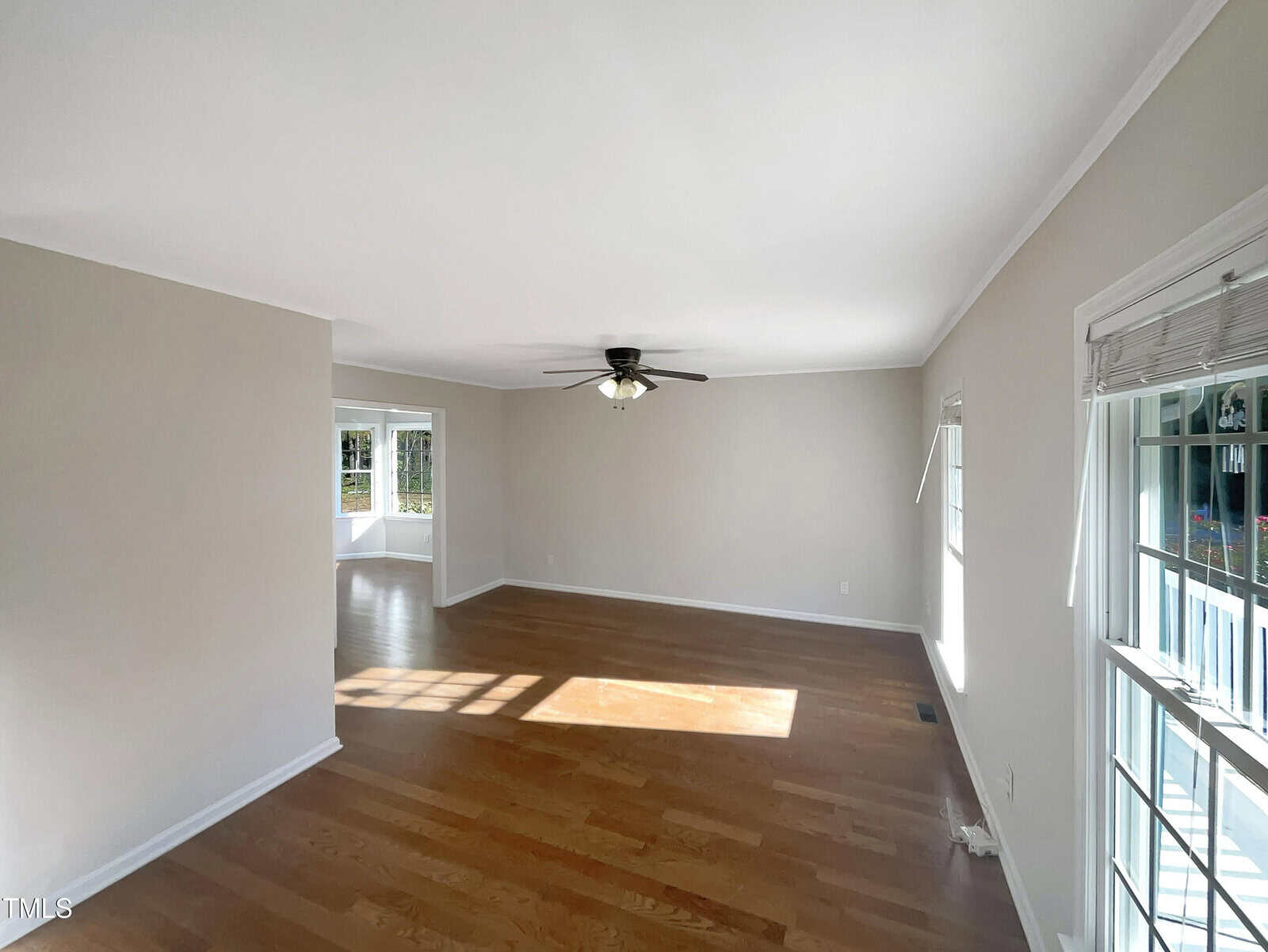 An empty living room with wooden floors and neutral-colored walls. A ceiling fan is centered above. Sunlight streams through multiple windows, creating patterns on the floor. A small, adjacent area is visible, separated by a corner wall.