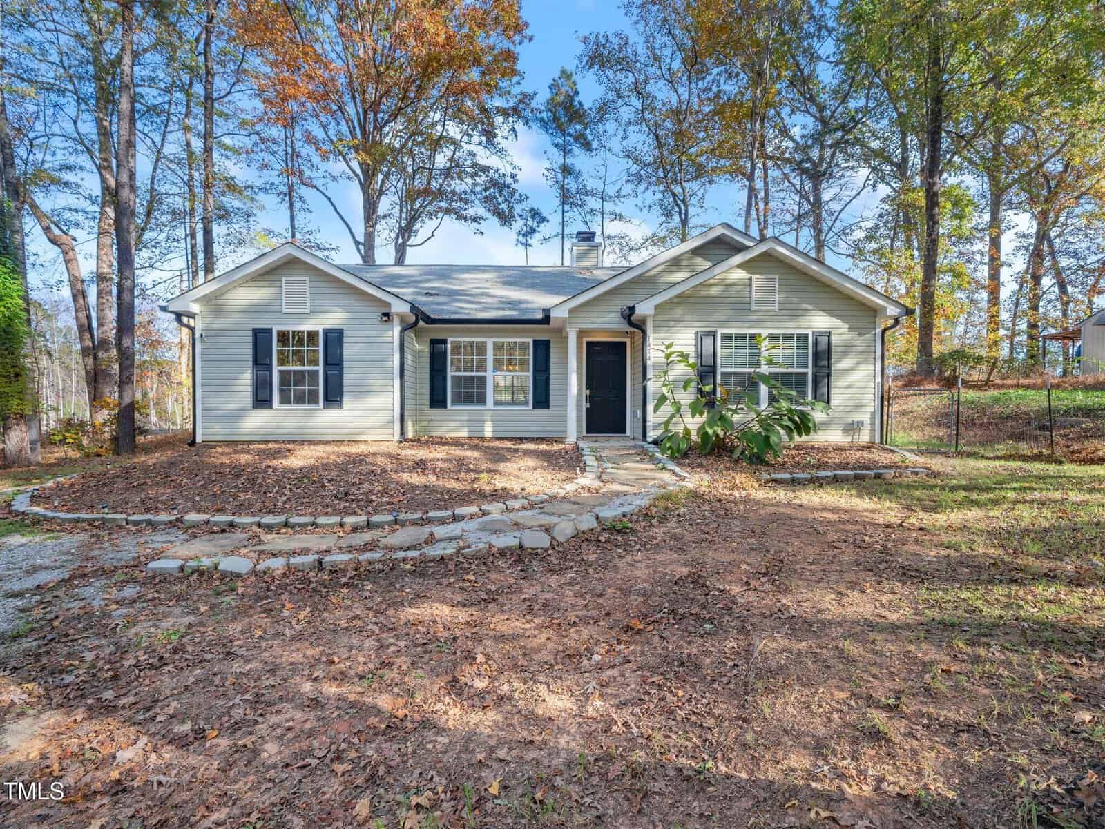 Located in Franklinton, this single-story house on 1414 Pocomoke 1 Drive features gray siding and black shutters nestled among tall trees. A paved stone path leads to a black front door, with a yard carpeted by mulch and fallen leaves, all under a clear blue sky.