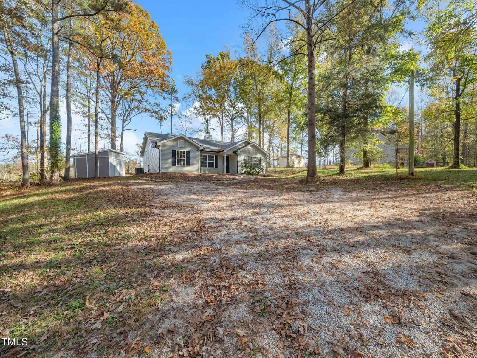 A single-story house with a light-colored exterior is surrounded by trees. There is a gravel driveway leading to the front door, and a small shed is visible to the left. The yard is covered in dry leaves, and the scene is lit by clear daylight.