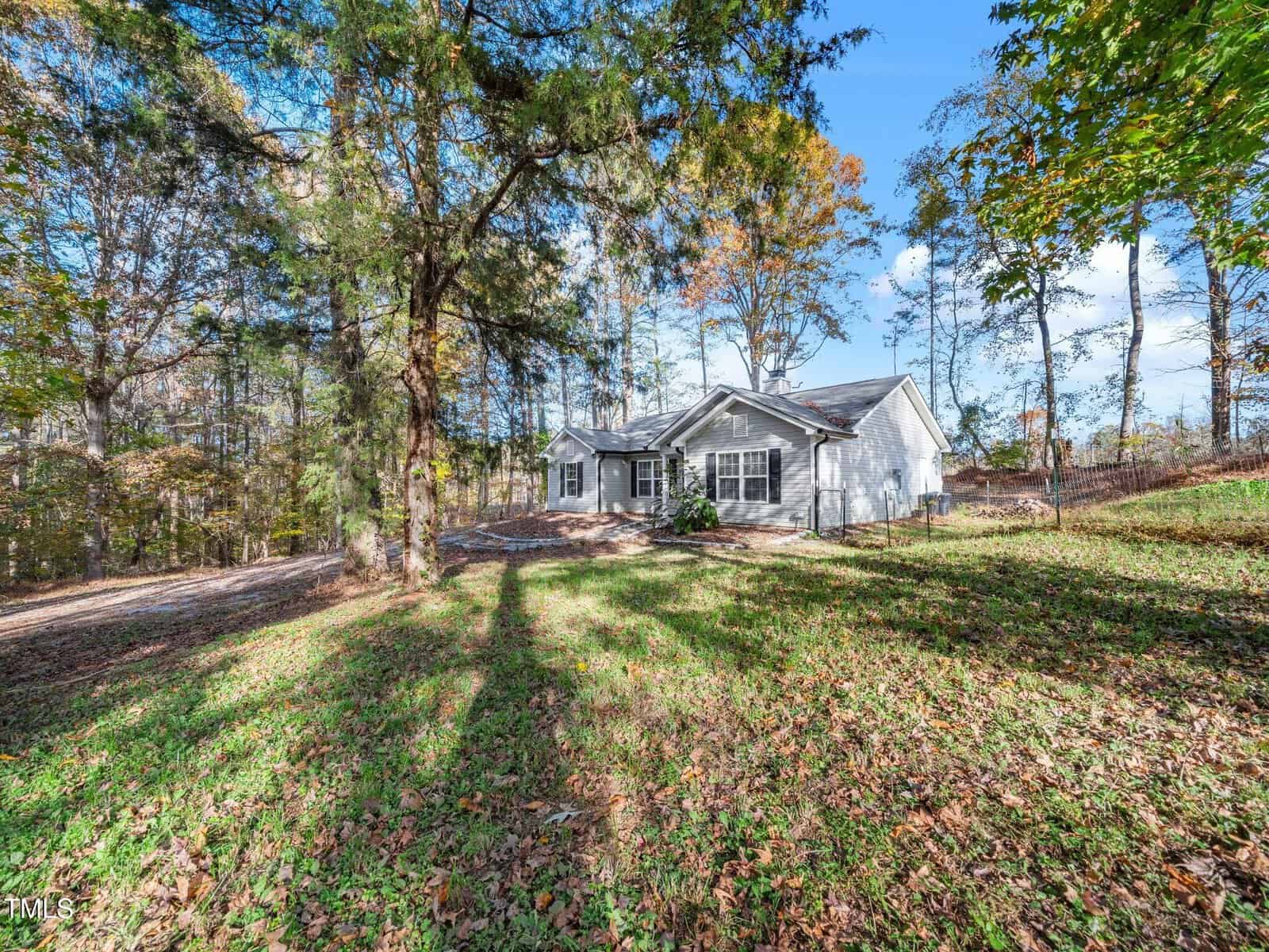 A ranch-style house with gray siding is situated on a grassy, wooded lot. The foreground includes scattered fallen leaves and tall trees, and the sky is clear and blue, indicating a sunny day.