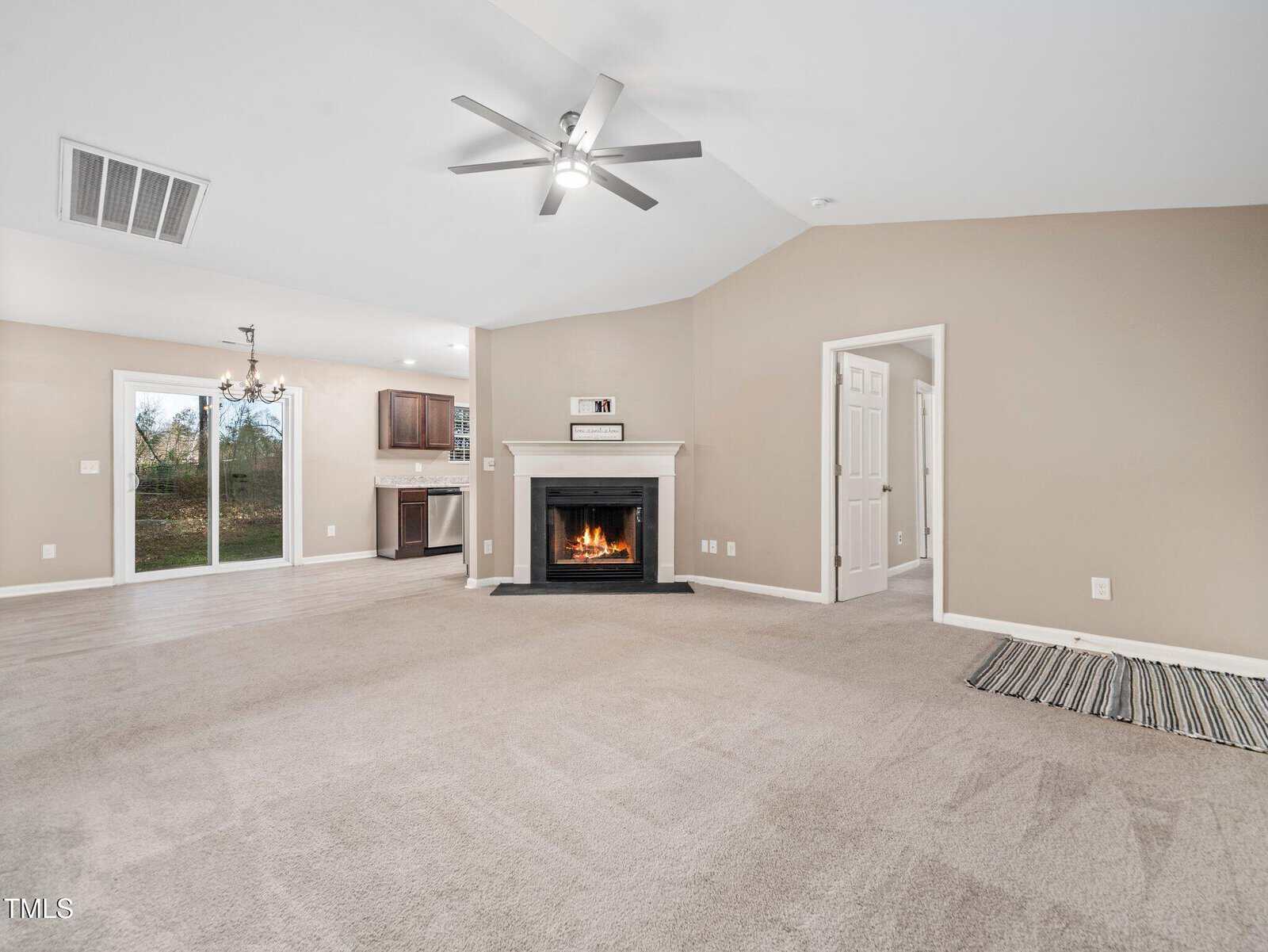 A spacious living room with beige walls and carpet features a lit fireplace with a white mantel. The room has a ceiling fan and a view into an adjacent kitchen area. Sliding glass doors lead outside, and a striped mat lies near a door.