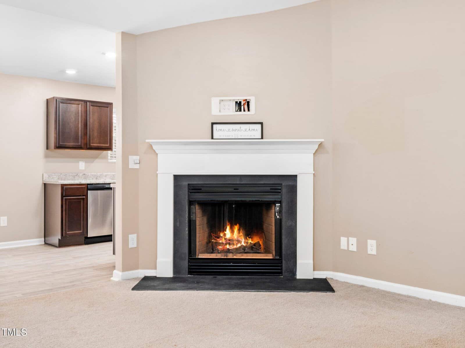 A living room features a lit fireplace with a white mantle. Above the mantle is a small decorative sign. The room has beige walls and carpeted flooring. To the left, part of a kitchen with dark wood cabinets and a stainless steel dishwasher is visible.