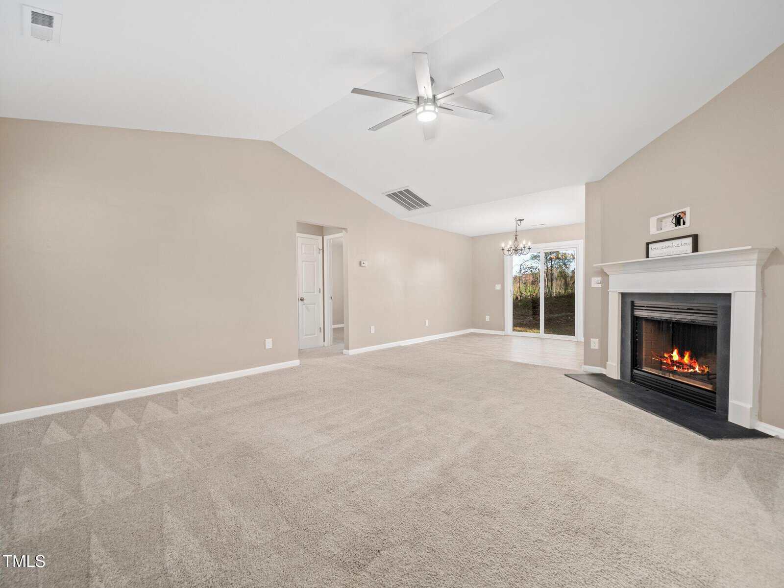 A spacious living room with beige walls and carpeted flooring. A lit fireplace is on the right side, and a ceiling fan is mounted above. A dining area is visible in the background, with a sliding glass door leading outside.