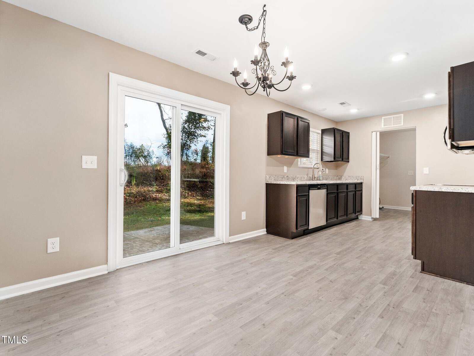 A spacious kitchen and dining area with light wood flooring. The room features dark wood cabinets, a stainless steel dishwasher, and a chandelier above. Sliding glass doors lead to an outdoor area. White walls and recessed lighting complete the space.
