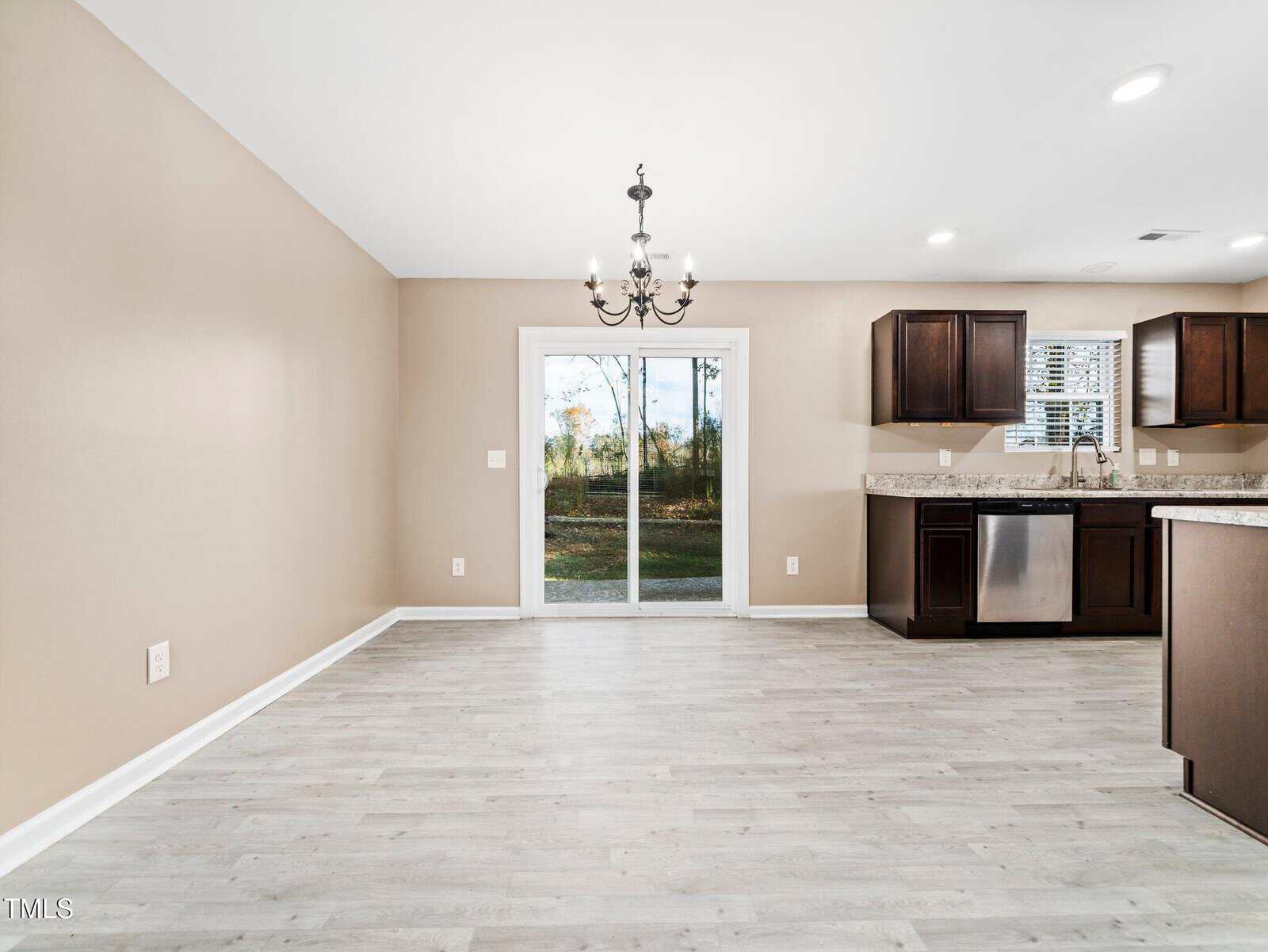 An empty room with light gray wood flooring, beige walls, and a dark chandelier. A sliding glass door leads outside. The kitchen area features dark cabinets, granite countertops, a dishwasher, and a window with white blinds.