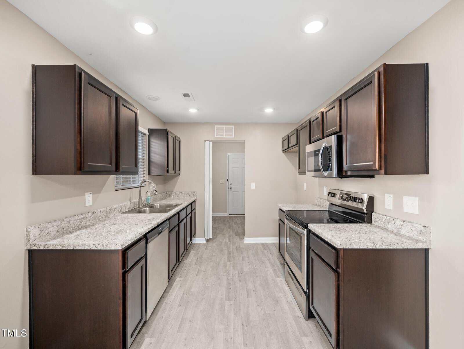 A modern kitchen featuring dark wood cabinets, granite countertops, and stainless steel appliances. The room has light wood flooring, recessed ceiling lights, and a sink by a small window. A door is visible at the end of the kitchen.