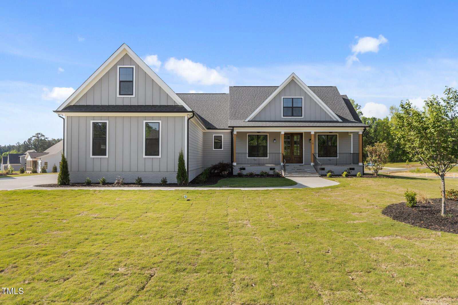 Single-story modern house with gray siding, white trim, and a dark shingled roof. The home sits on a large, neatly manicured lawn with young trees and minimal landscaping under a partly cloudy sky.