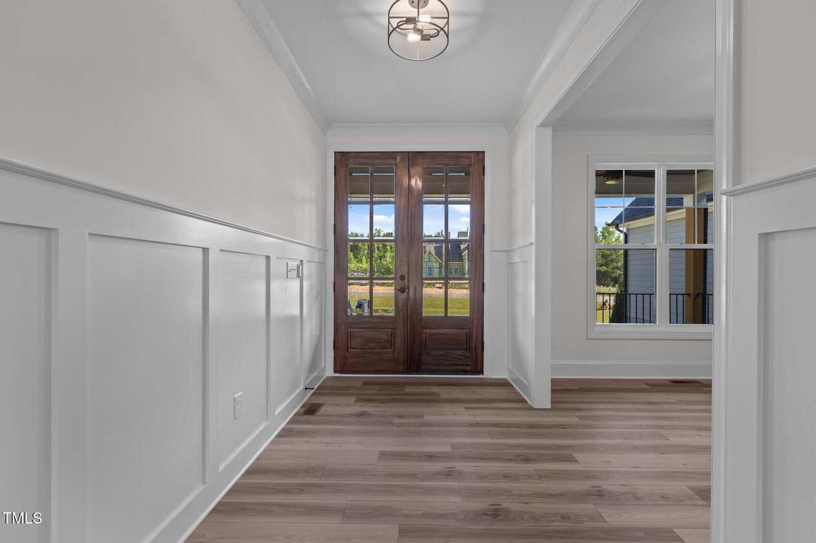 A hallway with light-colored wood flooring, white paneled walls, a modern ceiling light fixture, and double wooden front doors with glass panes. A window on the right reveals an outdoor view.