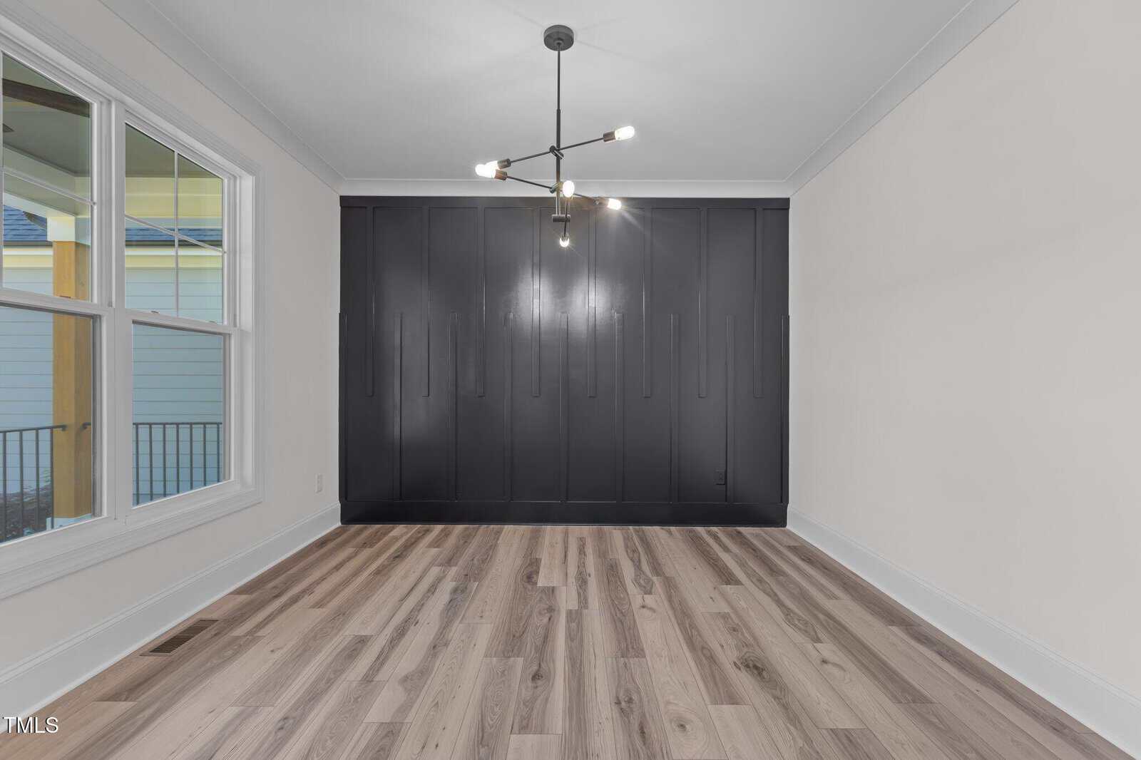 A modern, empty room with light wood flooring, white walls, a large black built-in wardrobe, a black chandelier, and a window showing an exterior view.