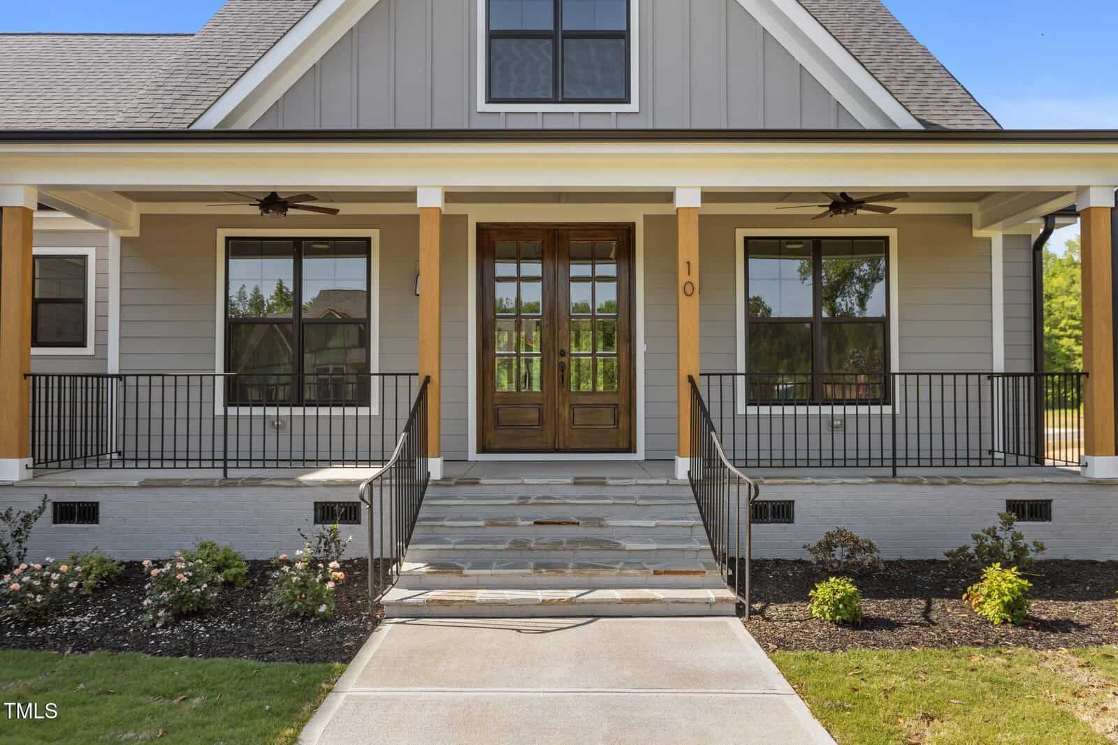 A modern single-story house with a covered front porch, wooden double doors, two large front windows, black railings, stone steps, and landscaped shrubs along the walkway.