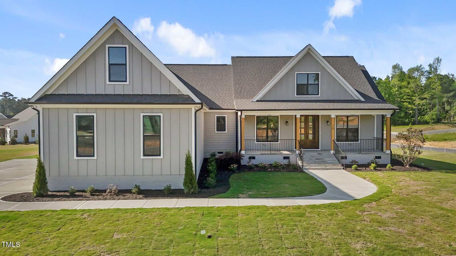 A single-story house with beige siding, dark roof shingles, and a covered front porch with railings. The lawn is partially green with a concrete walkway leading to the front steps. Trees and a road are in the background.