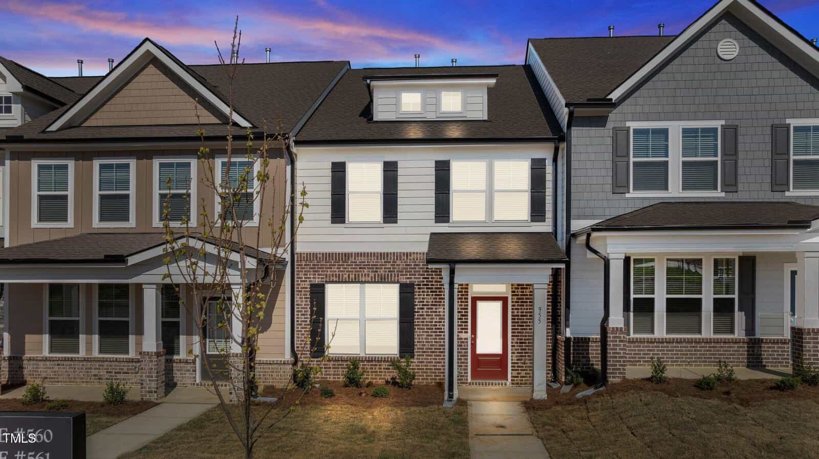 A two-story townhouse at 955 Alma Railway Drive, Wake Forest, features brick and siding exterior, white-framed windows, and a red front door. Captured at sunset with a purple sky, a small tree and walkway lead to the welcoming entrance.