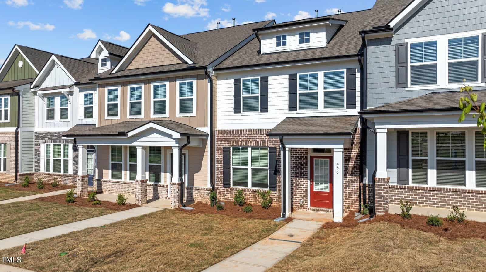 A row of modern two-story townhouses with brick and siding exteriors, large windows, and small front yards with young shrubs and pathways leading to each entrance.