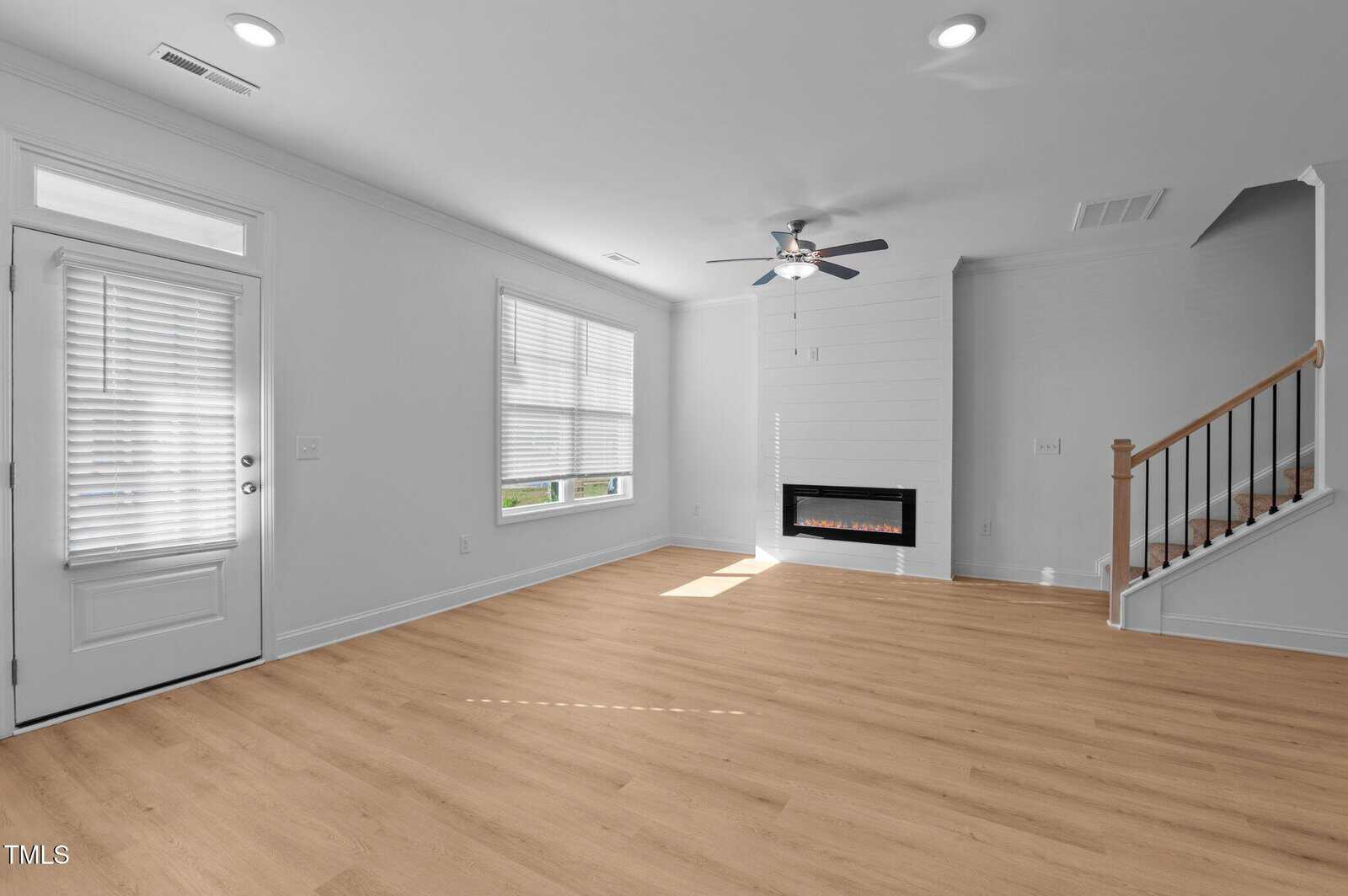 A bright, empty living room with light wood flooring, white walls, large windows with blinds, a ceiling fan, a modern fireplace, and a staircase with a wooden handrail.