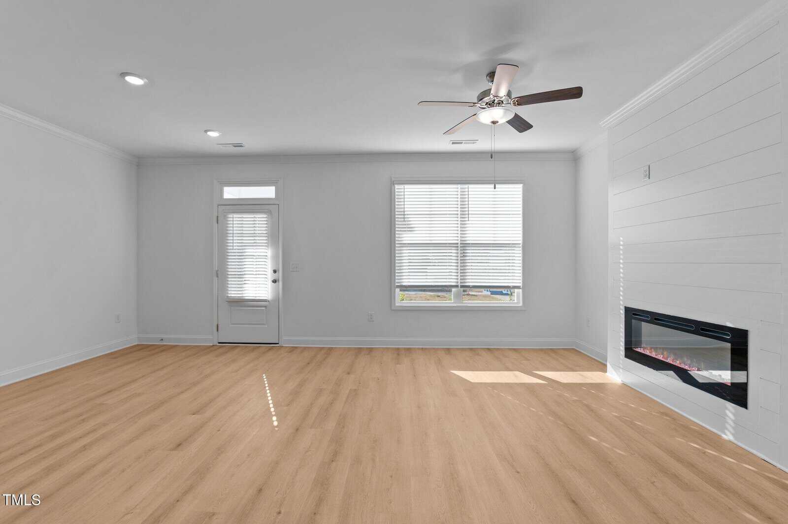 A bright, empty living room with light wood flooring, white walls, large window, glass door, ceiling fan, and a modern electric fireplace on the right wall.